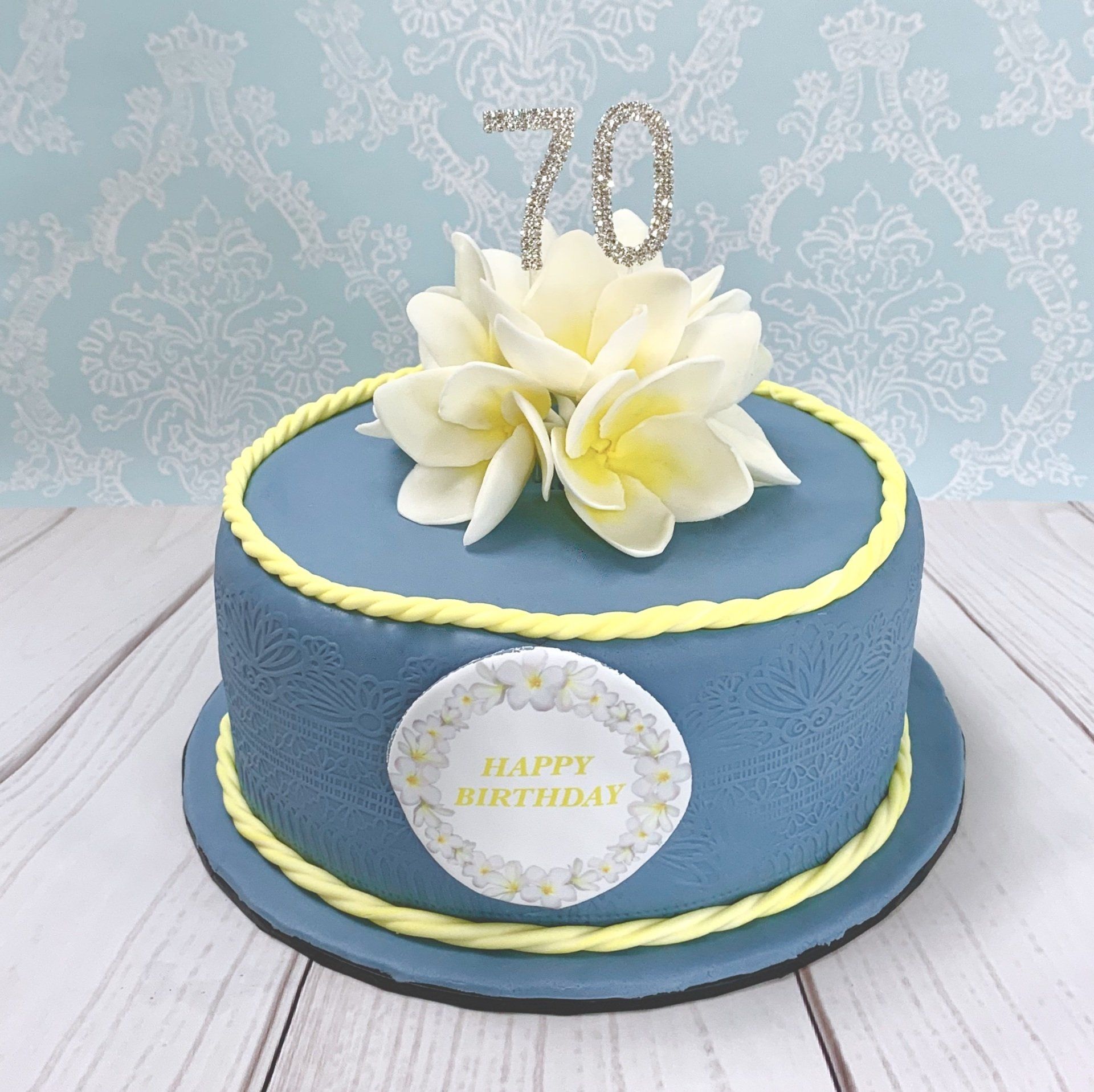 Soft Blue Embossed Birthday cake with soft yellow trim. A Diamante 70, Handmade sugar frangipani flowers, and an edible frangipani happy birthday plaque.