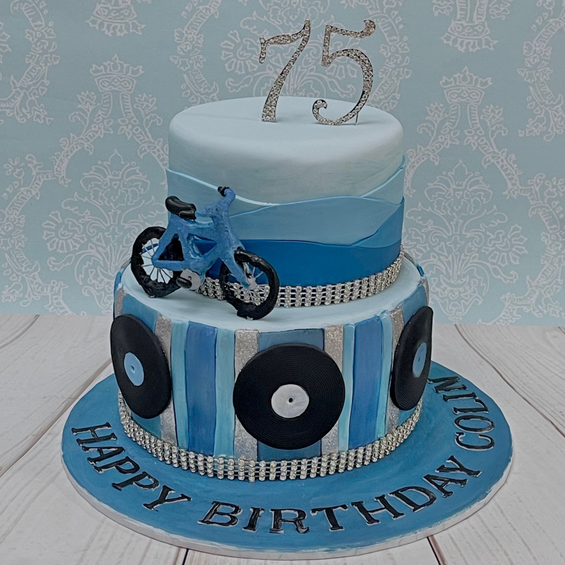 Two tier blue and silver themed cake with Diamante topper, diamante trim and hand made bicycle on the top tier with fondant mountains and the base tier Fondant blue and silver stripes with fondant vinyl records.
