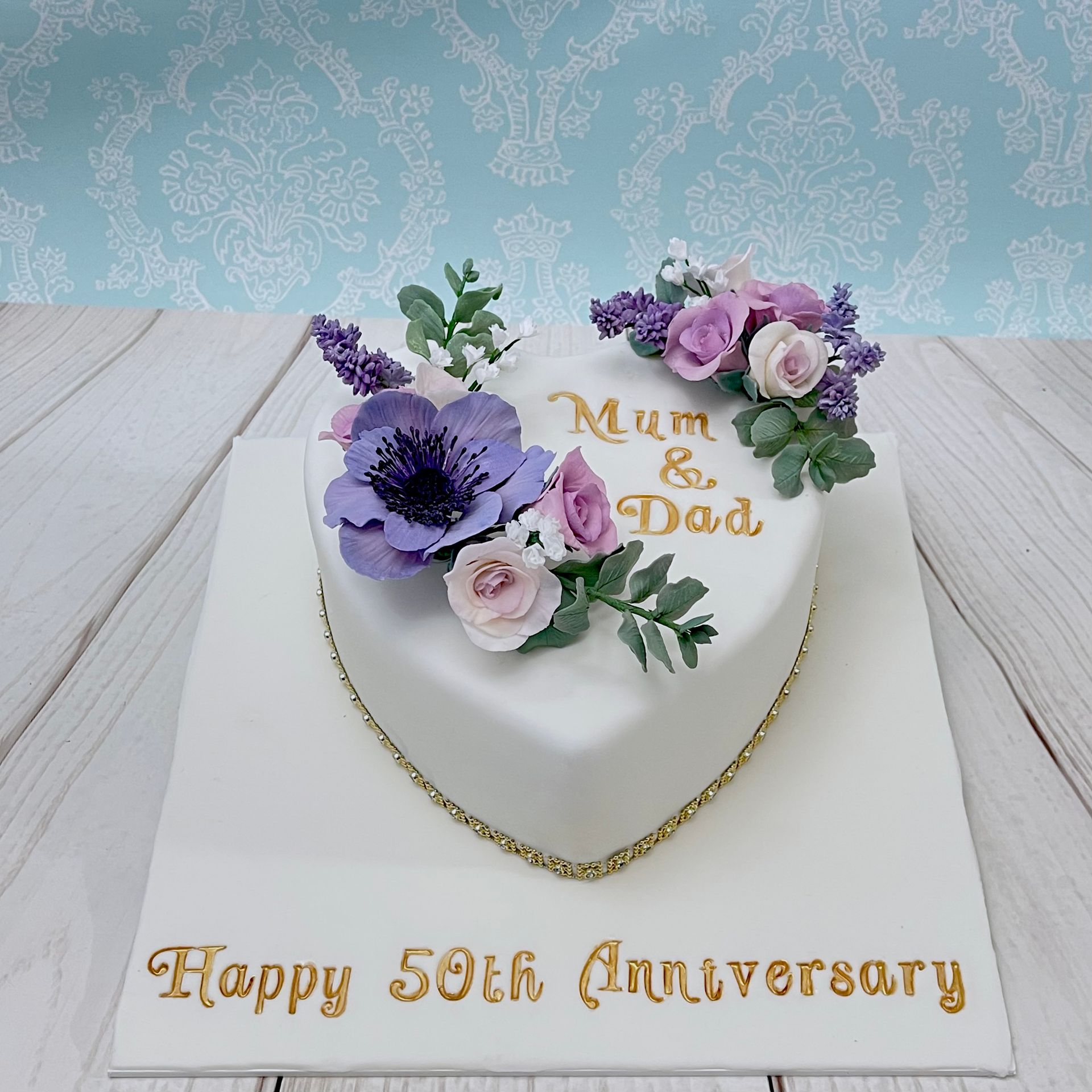 50th Golden Anniversary heart shaped cake. White fondant. Mauve Sugar Anemone Flower, Sugar Eucalyptus Leaves, Sugar Lavender, Sugar roses and wafer paper baby's breath, . Hand painted Gold sentiments and Gold Diamanté trim around the base.