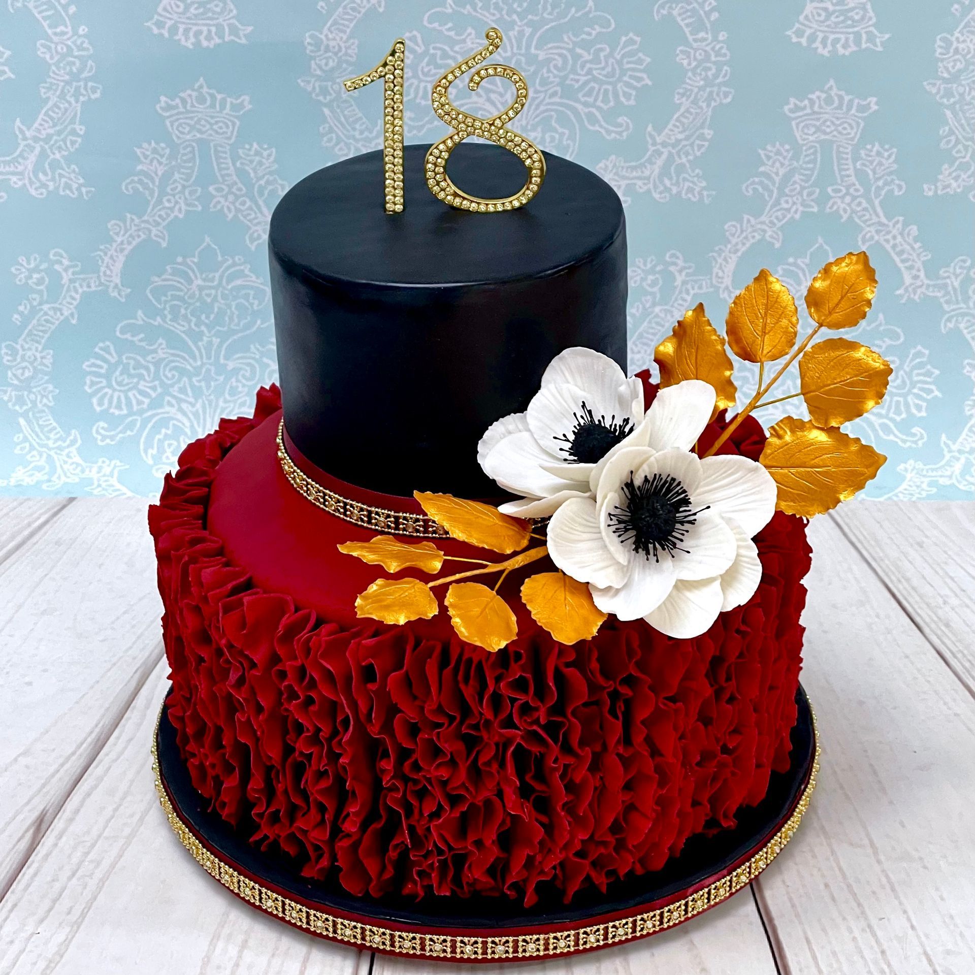 Glamerous 18th Birthday Cake, Base tier red ruffles, top tier black shimmer, with White sugar aneomone flowers, gold sugar leaves, and a gold diamante 18 and trim.