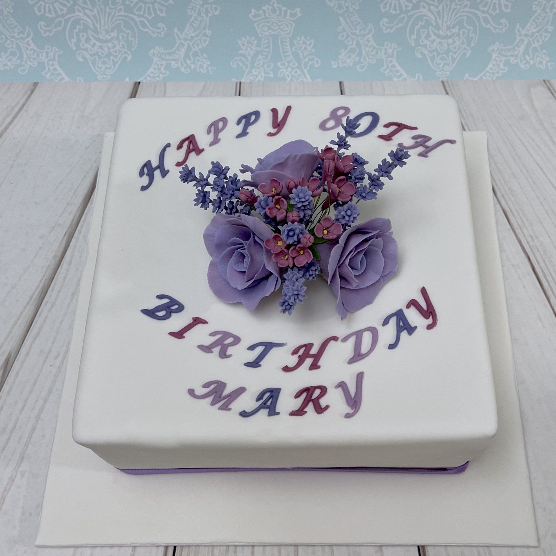 80th Birthday cake with white fondant, mauve sugar roses, sugar lavender flowers and sugar lilac flowers, with purple writting.