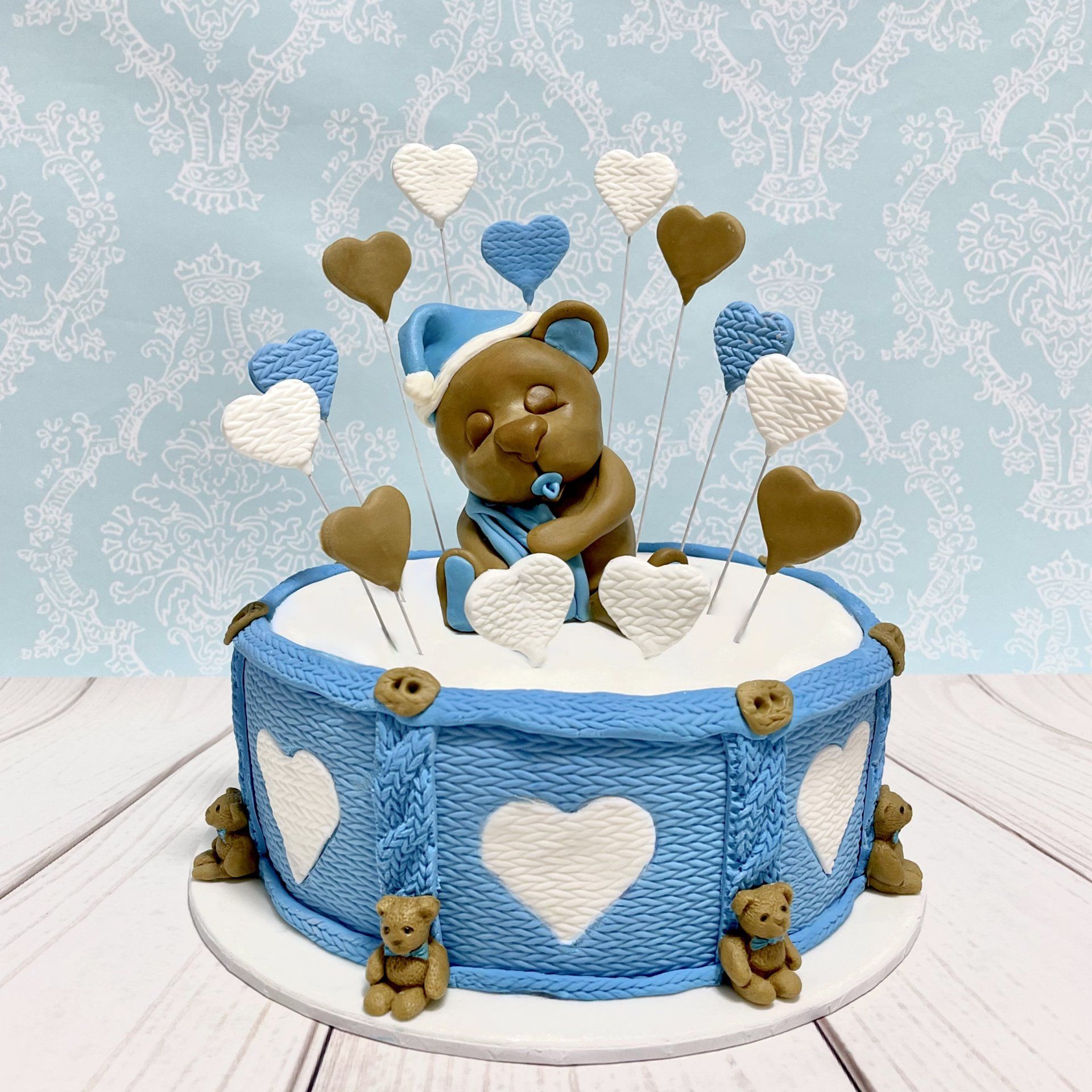 Boys Baby Shower Cake. White fondant with a blue and white knitted effect around the sides, Fondant Hearts and a cute hand sculped fondant sleepy baby bear with his Dummy, Blanket  and Bedtime hat. And more miniature bears around the board
