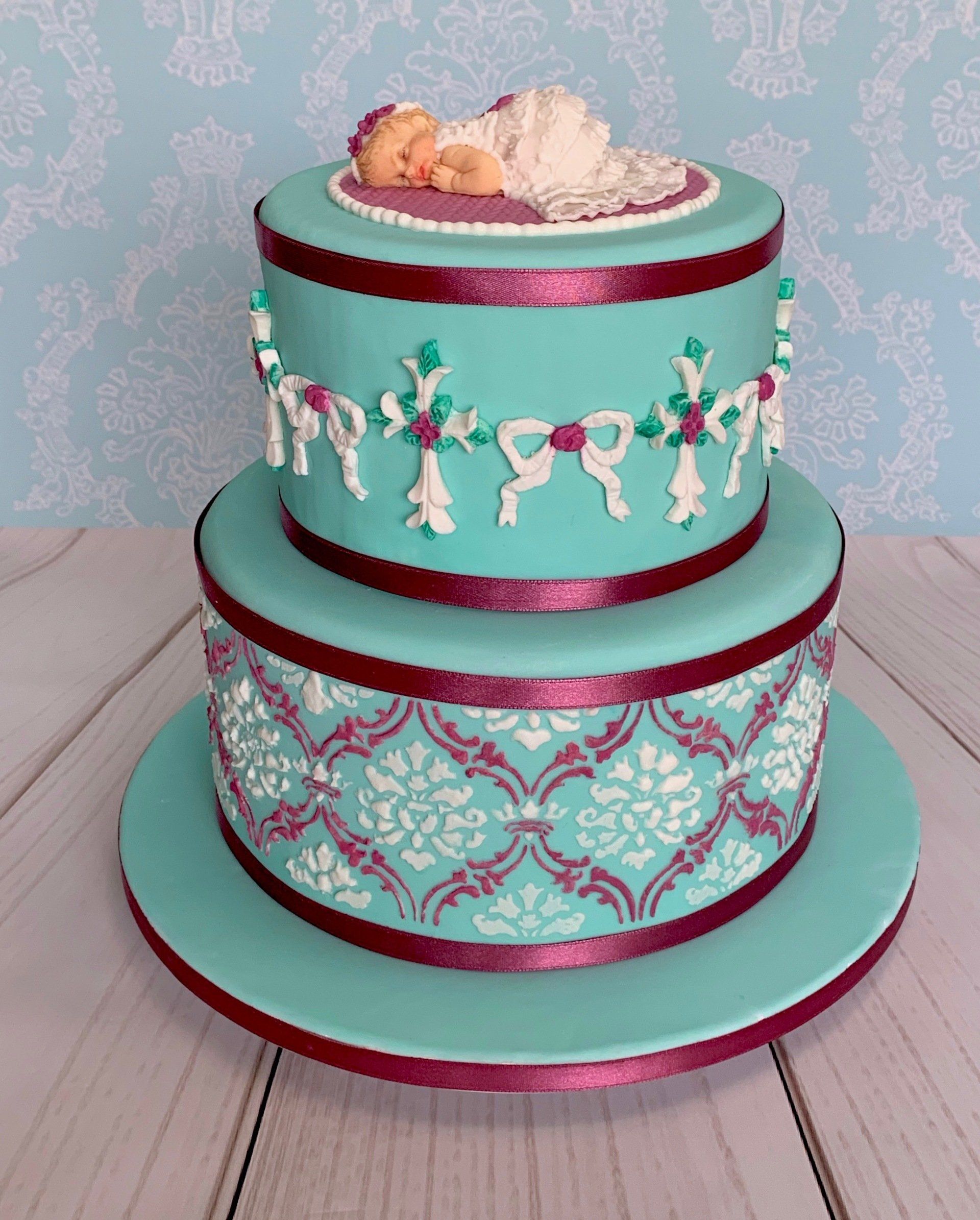 Two tier Christening cake in Aqua, white and pink with stenciling, crosses, bows and a fondant baby in a fondant christening gown.