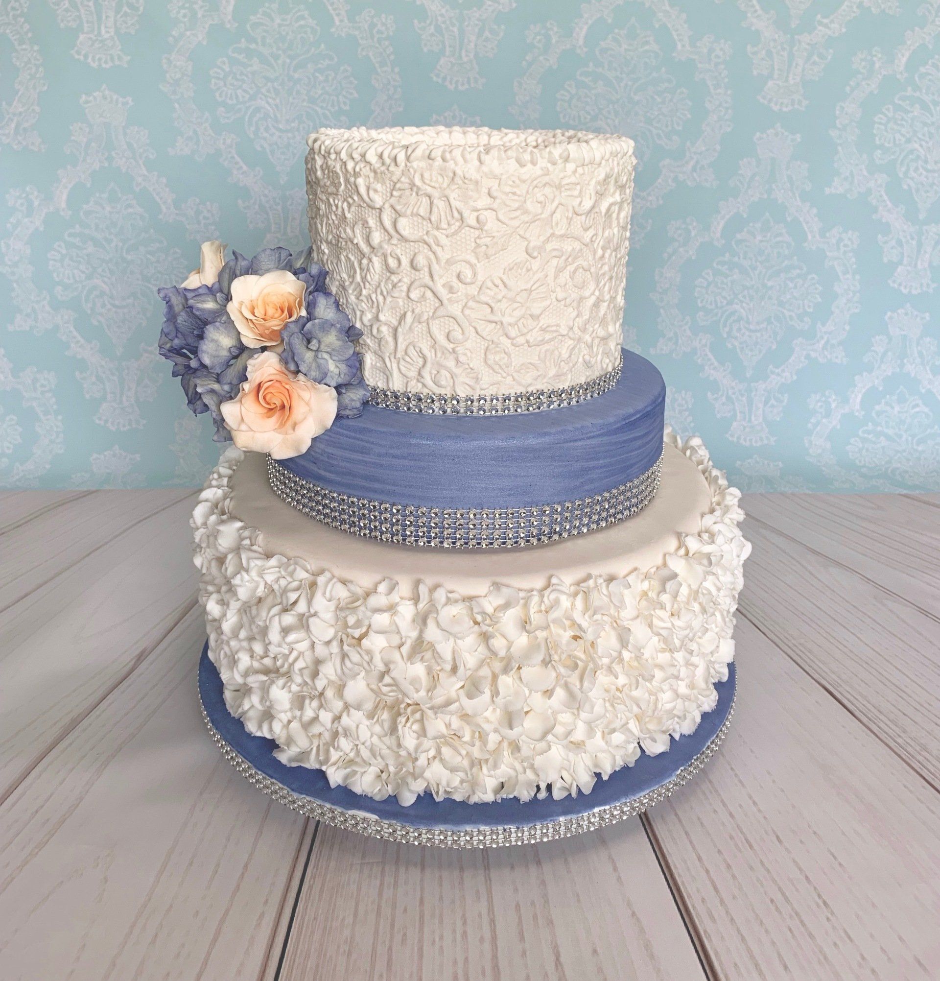 Three tiered wedding cake, in white and cornflower blue. Ruffle base, piped lace top with sugar roses and hydrangeas.