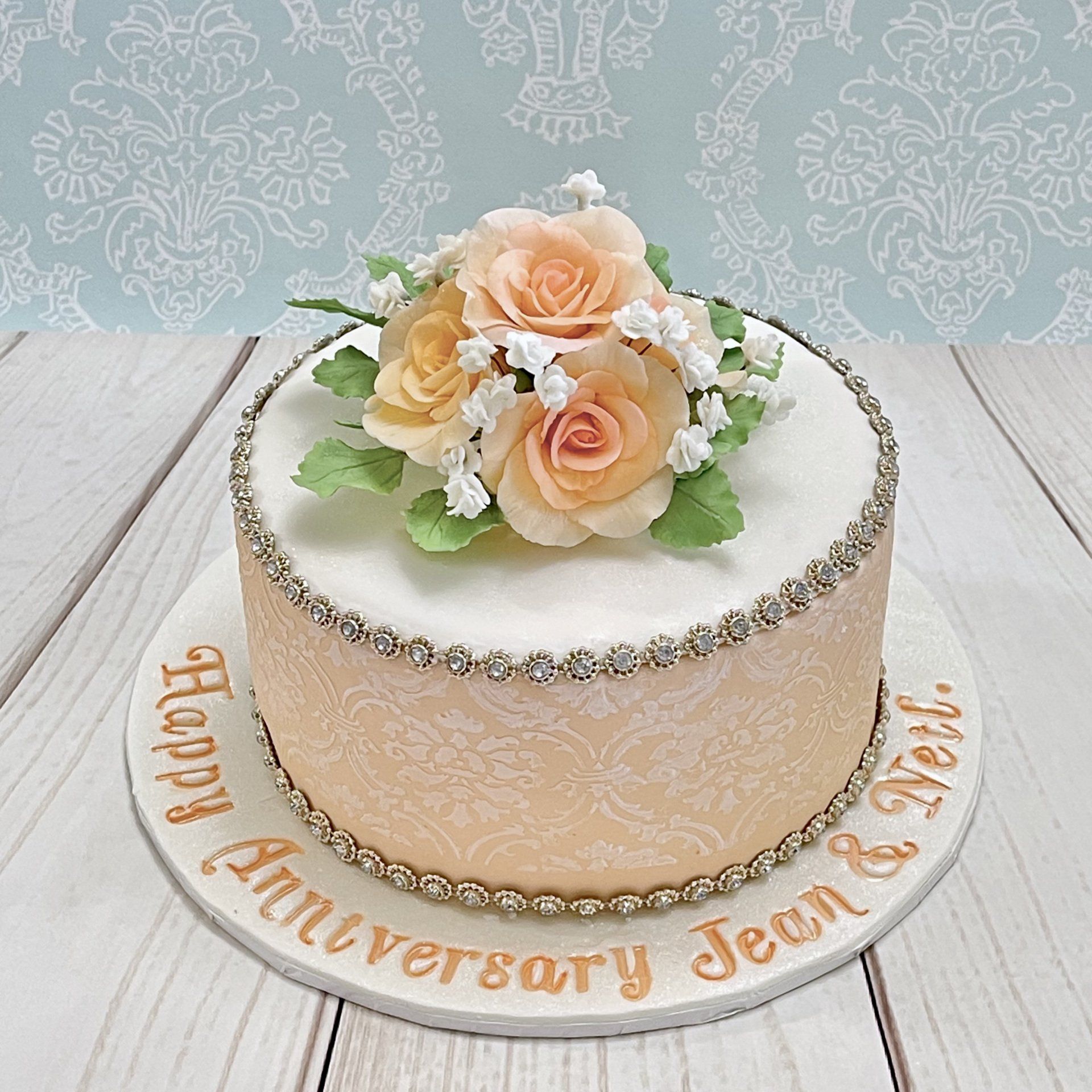 60th Diamond Wedding Anniversary cake. Pale apricot fondant with white damask stenciling around the sides and a diamante trim at the top and bottom of the sides too. Top white sparkle fondant with a posy of soft apricot sugar roses, white sugar baby's breath flowers, and soft green sugar leaves. Board white sparkle fondant with hand painted soft apricot sentiments.