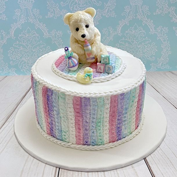 Gorgeous neutal Baby Shower Cake. White fondant with a blue, pink, aqua, mauve and white crochet effect around the sides, Fondant crocheted rug on the top, with a cute hand sculped fondant baby bear holding a bottle, and surrounded by moulded fondant baby toys.