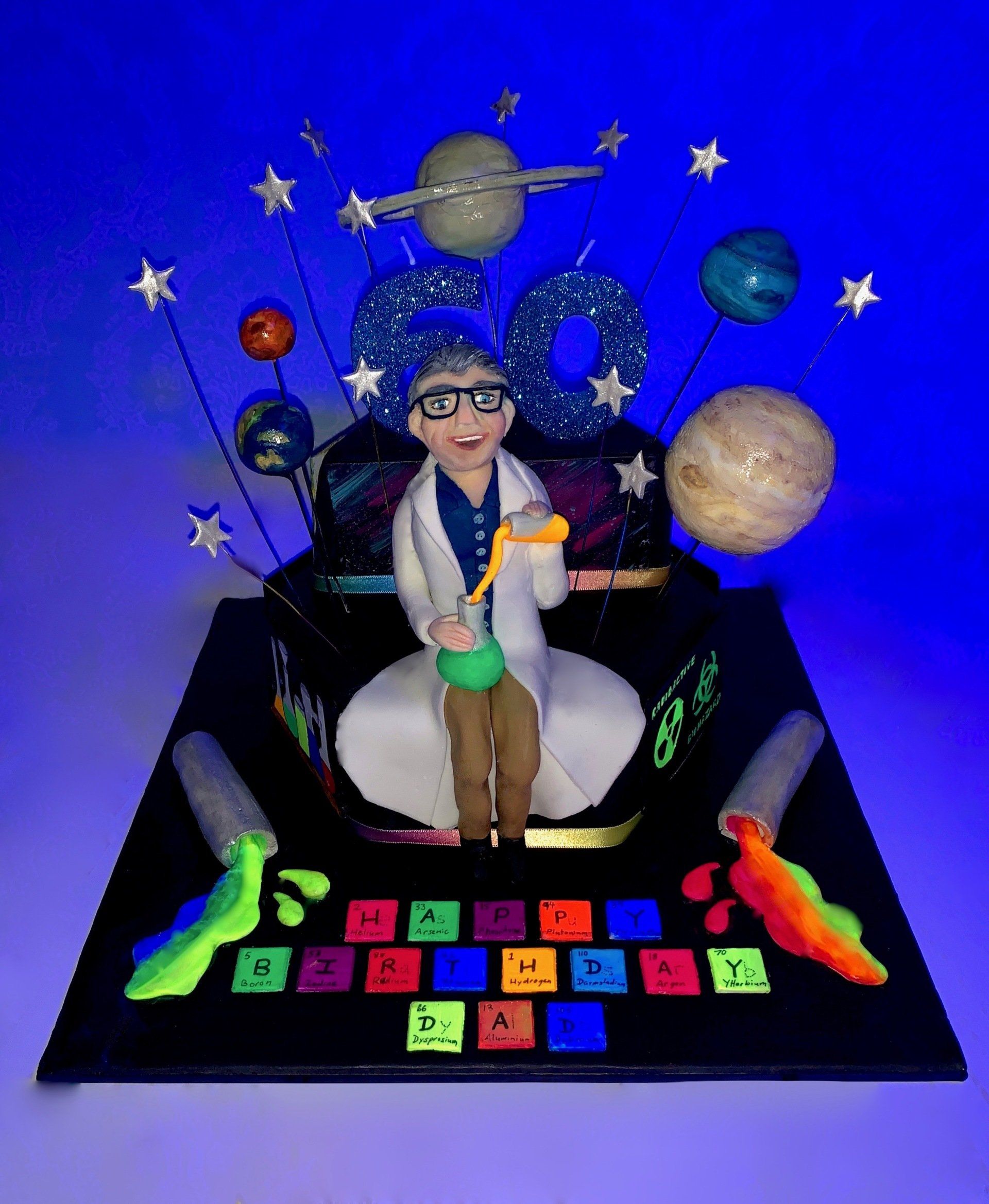 A tiered hexagonalm, Science themed cake with Planets, A mad scientist figure, and lots of other science related details. Front, glowing under a black light.