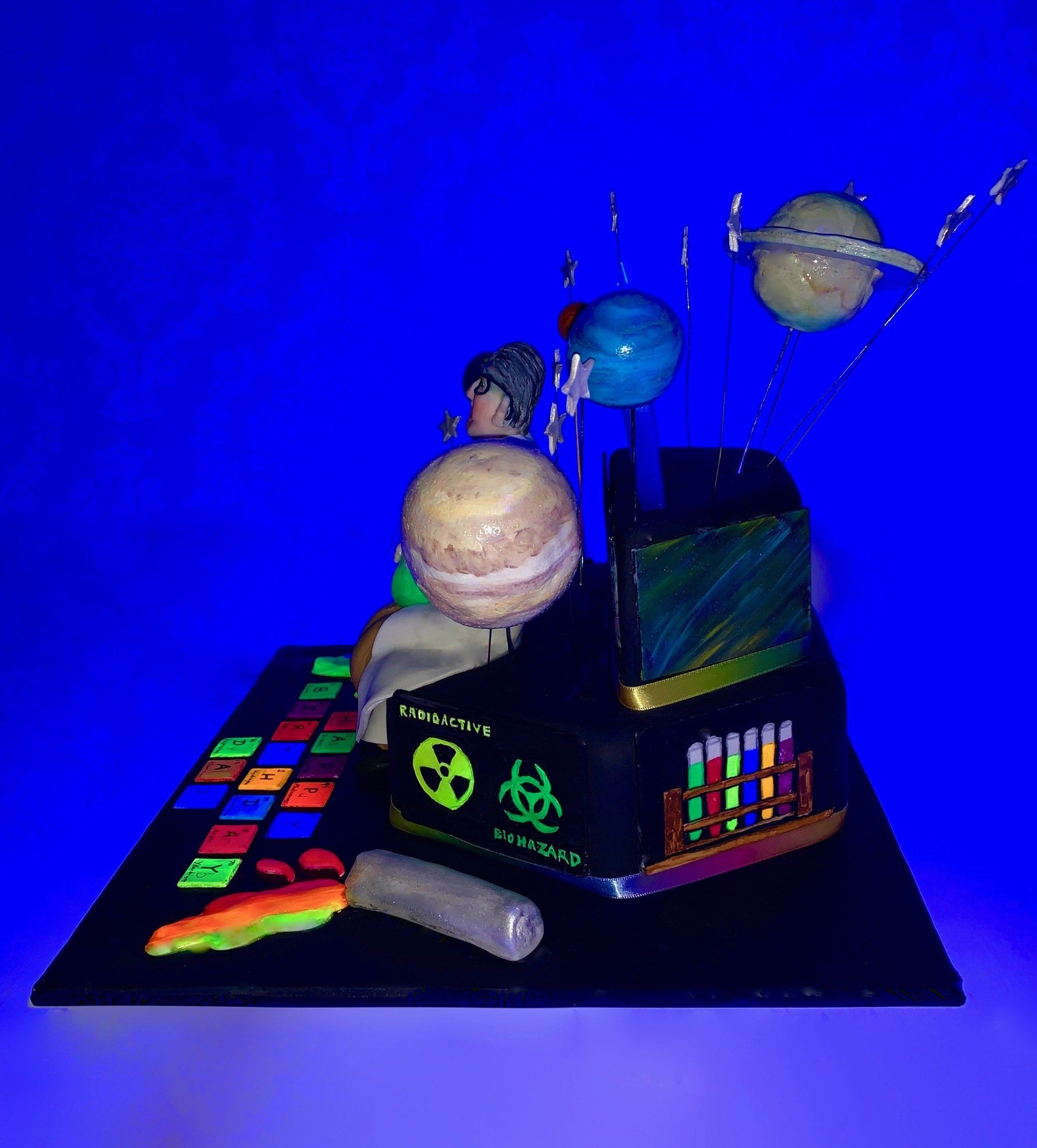 A tiered hexagonalm, Science themed cake with Planets, A mad scientist figure, and lots of other science related details. Side 2, glowing under a black light.