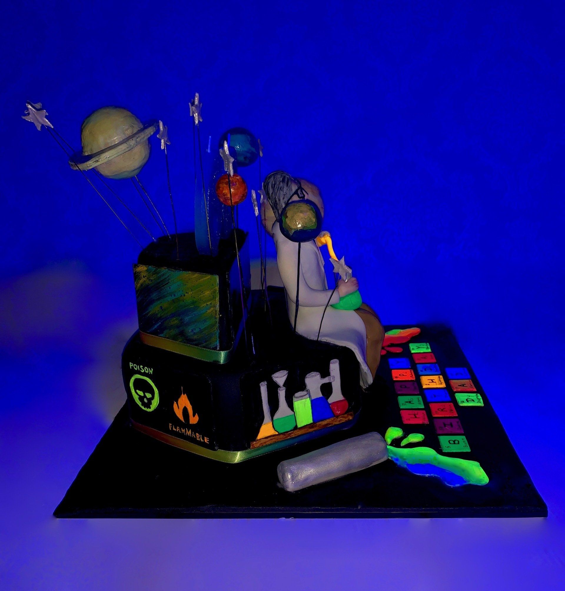 A tiered hexagonalm, Science themed cake with Planets, A mad scientist figure, and lots of other science related details. Side 1, glowing under a black light.