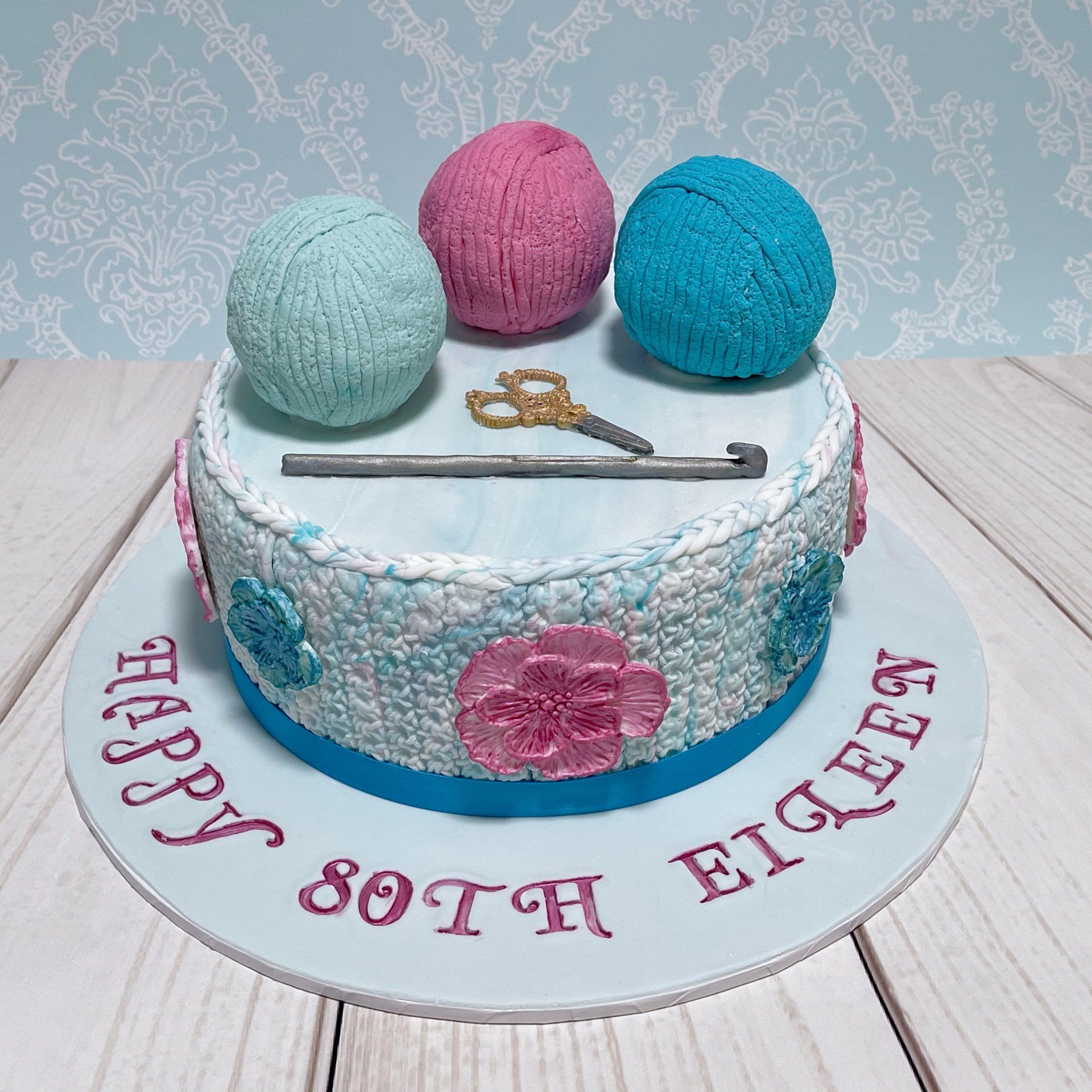 80th Crochet Themed Birthday Cake. With fondant Balls of Wool, Fondant Scissiors, Fondant Crochet hook and Fondant Crochet texture around the sides. In Aqua, Teal and Pink Colours.
