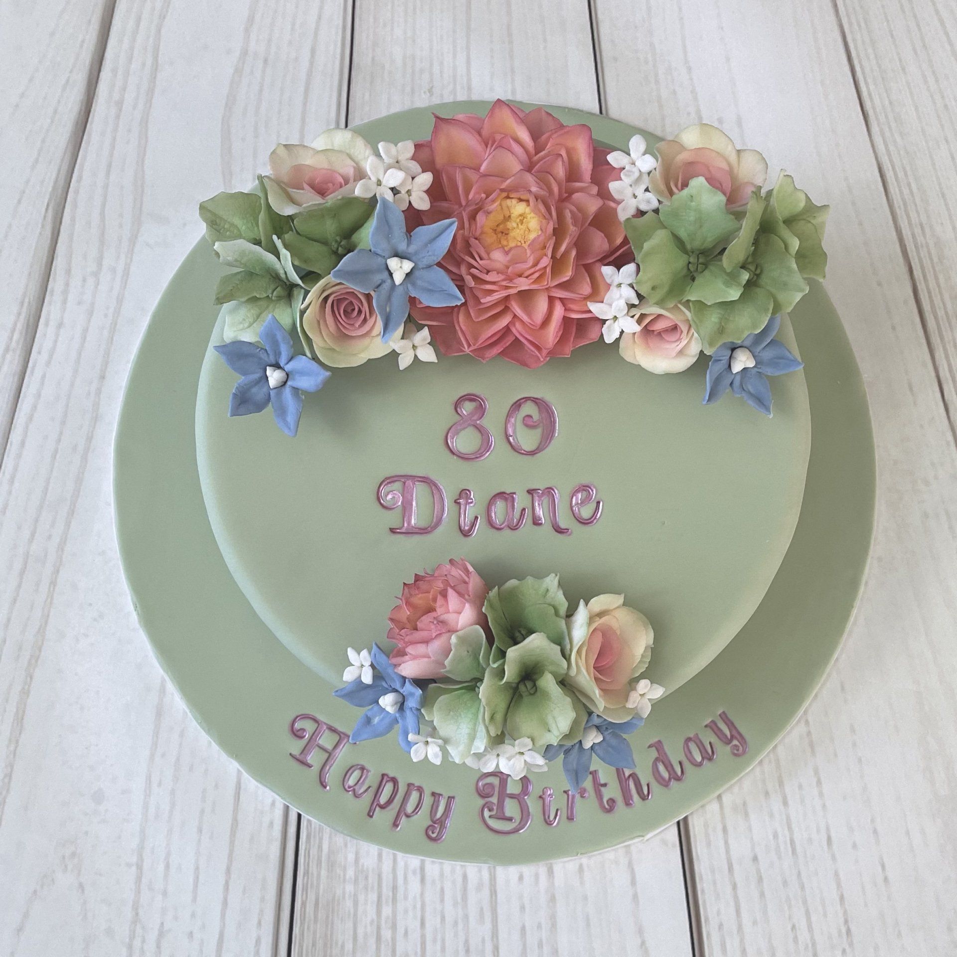 Soft Green 80th Birthday Cake with pastel pink sugar dahlias, sugar roses in white and pink, sugar bluebells, soft green sugar hydrangeas and white sugar filler flowers. finished with hand painted pink writing.