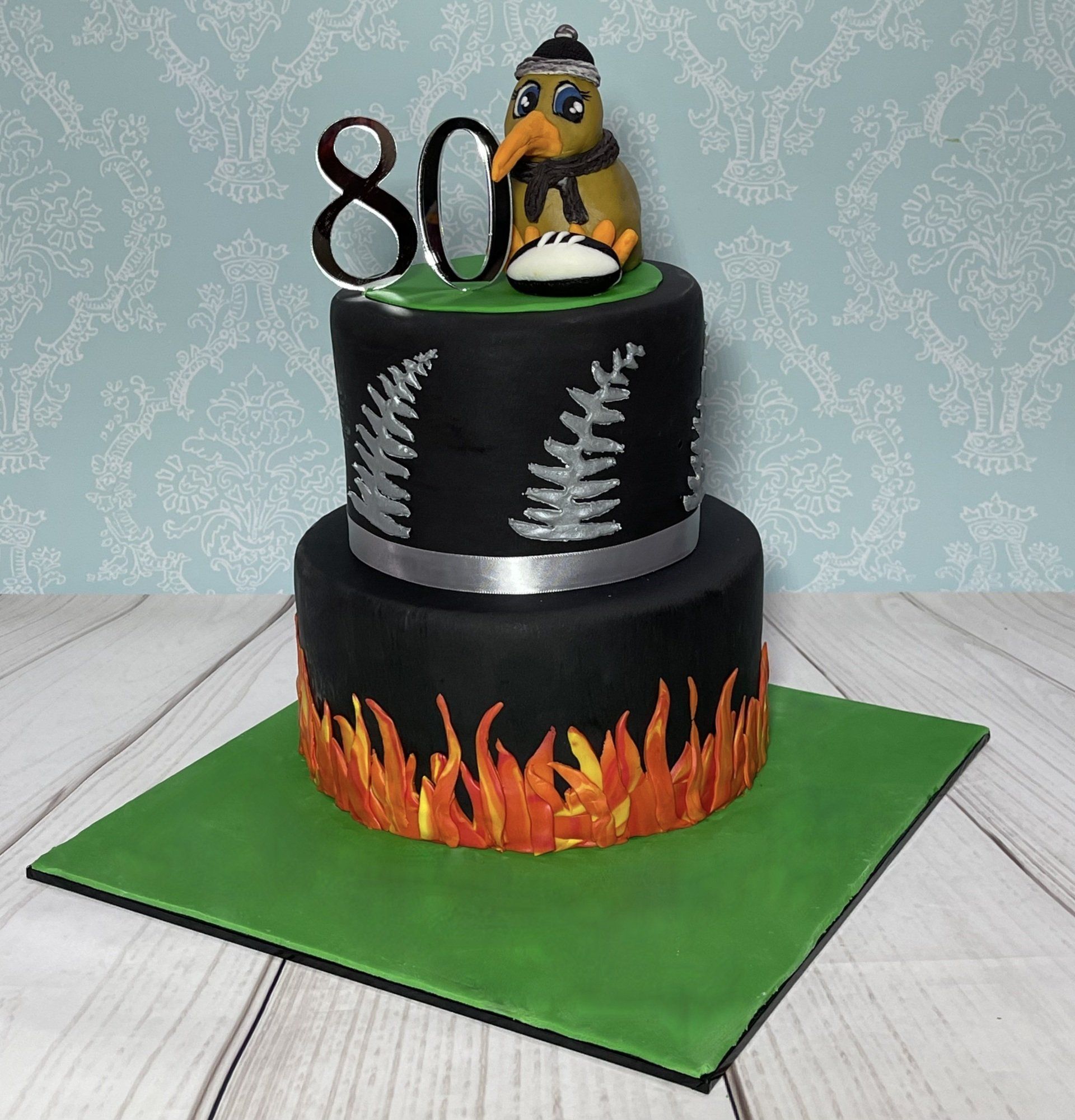 Two tier cake covered with black fondant, with a hand sculpted kiwi bird, a foot ball, and fern leaves on the top tier and flames on the base tier.