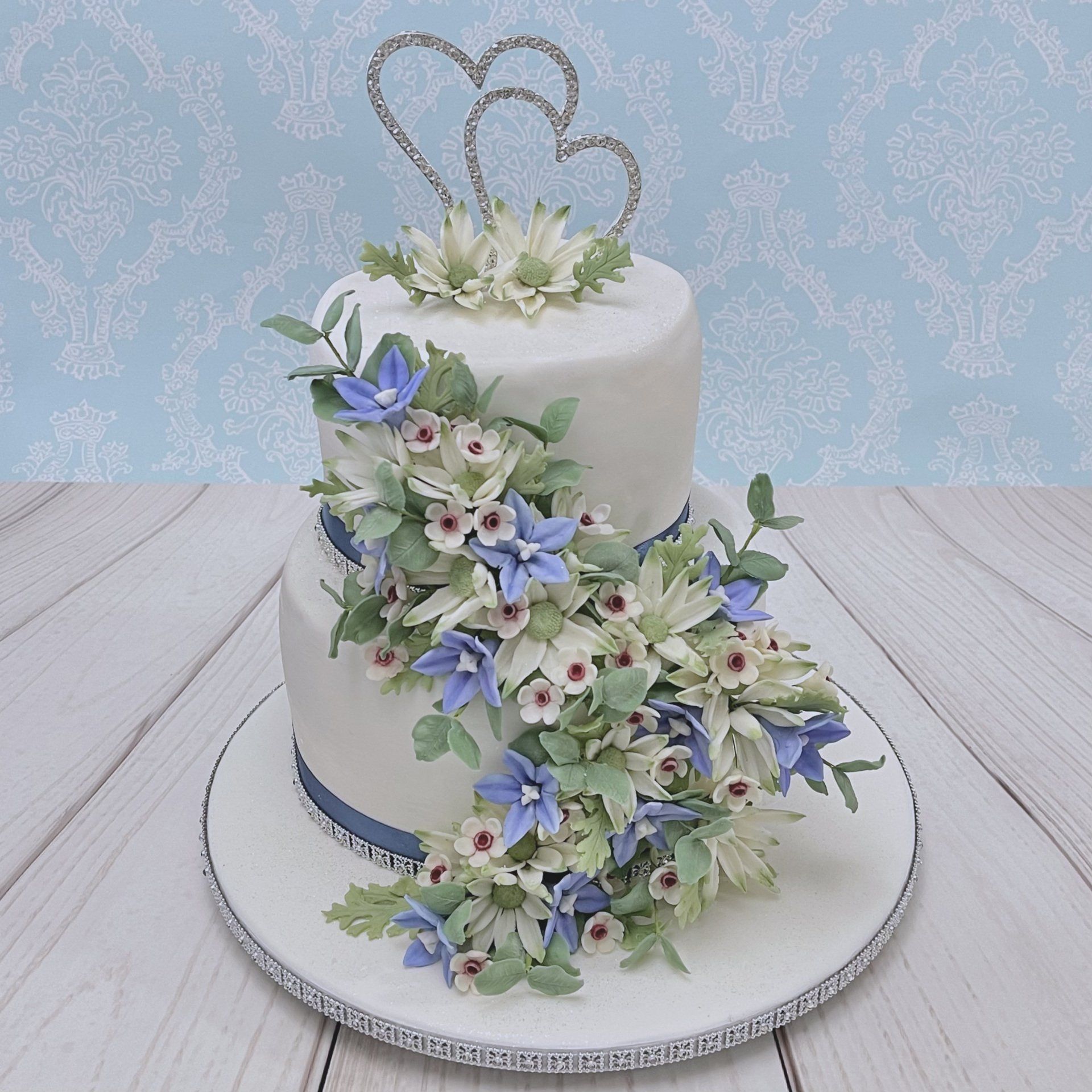 Two tier Wedding cake. White fondant with dusty blue ribbon and diamanté trim and a cascade of Australian Native sugar flowers including Flannel Flowers, Australian blue bells, Geraldton wax, and eucalyptus leaves. Finished with a Diamanté double heart topper.