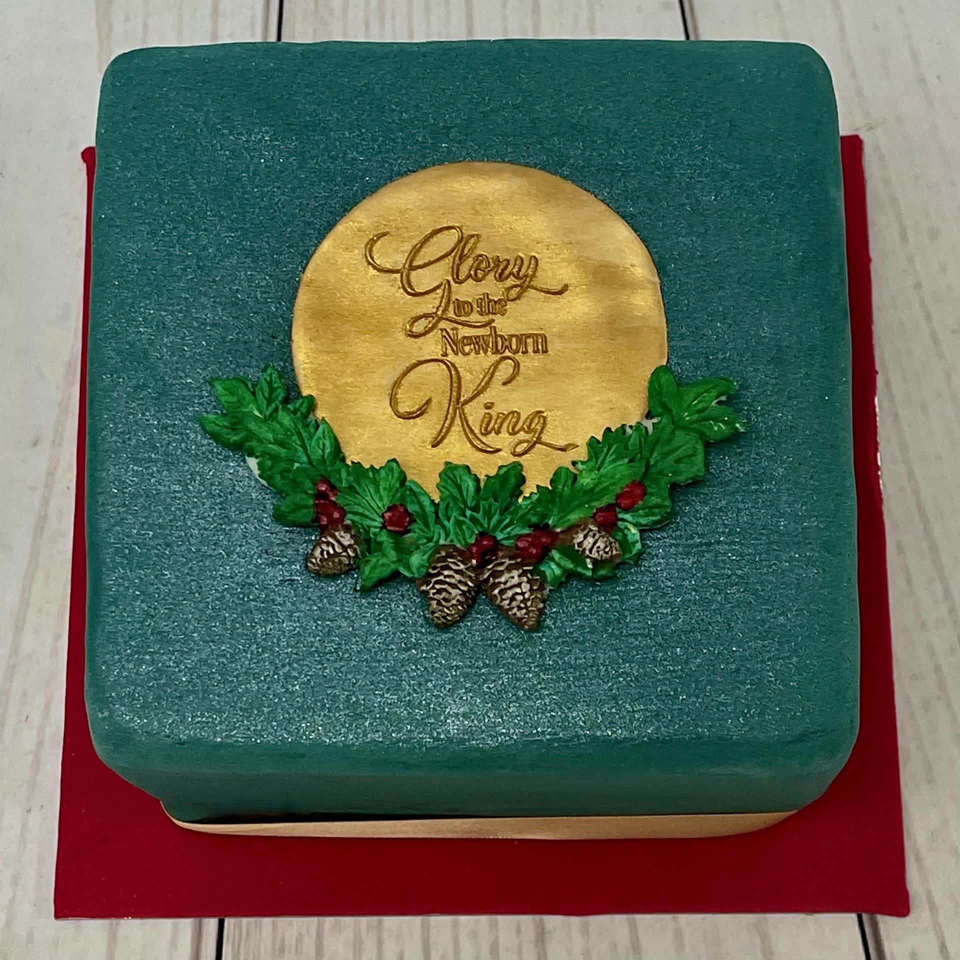 Christmas Cake in Teal green and red fondant. fondant holly and pine cones and a gold fondant plaque that says Glory to the new born King.