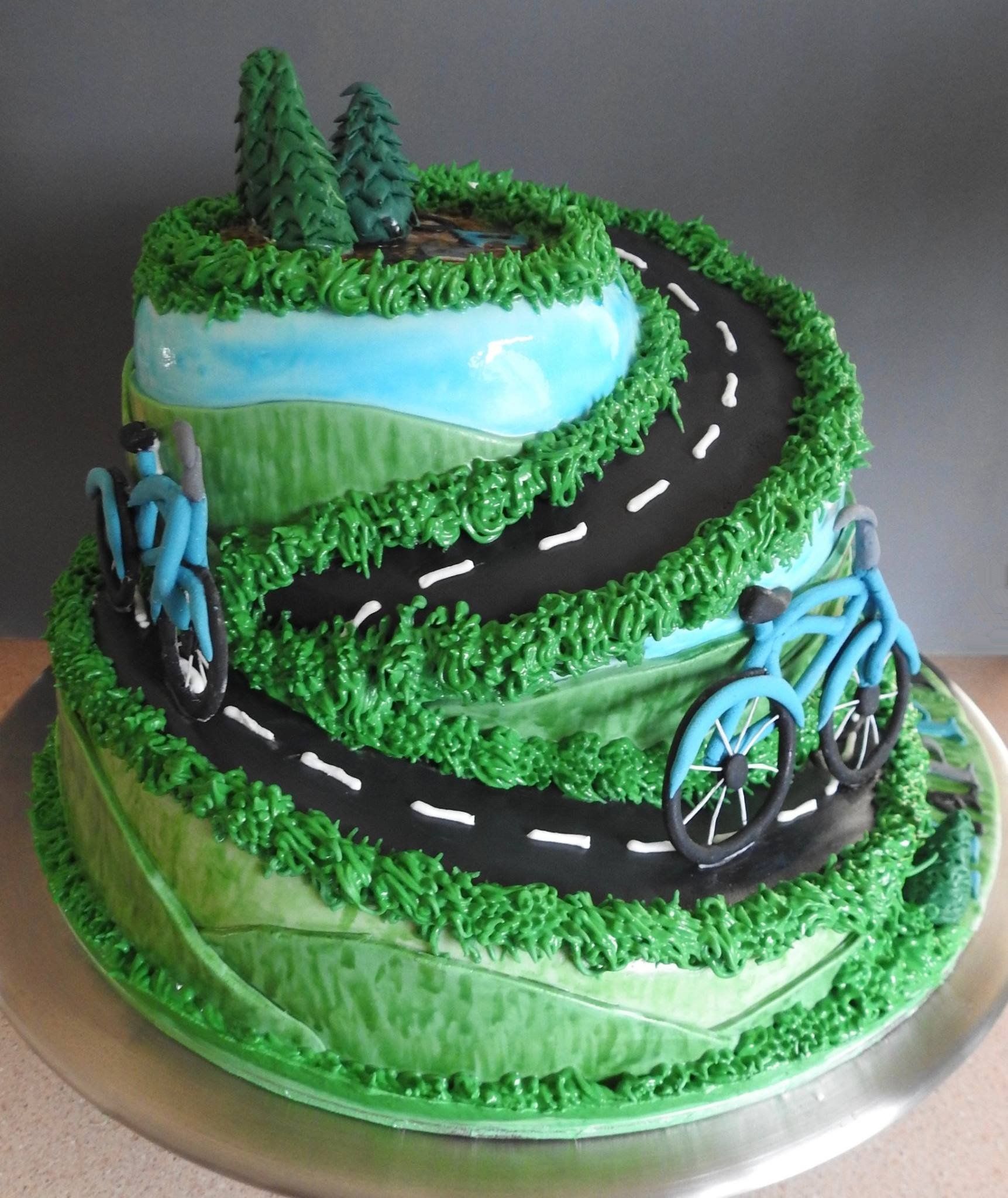 Tierd and shaped birthday cake with fondant hills, trees and two bicycles coming down a road.