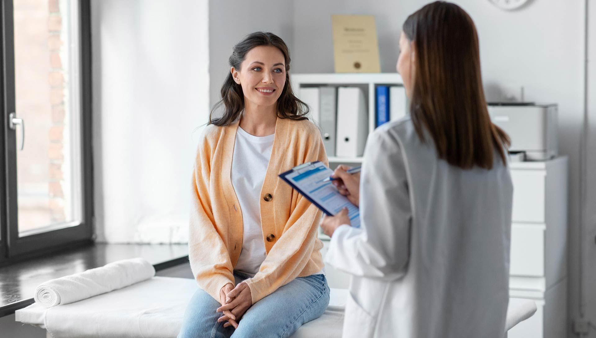 A patient sits on an exam table while a medical professional in a white coat takes notes on a clipboard in an office.