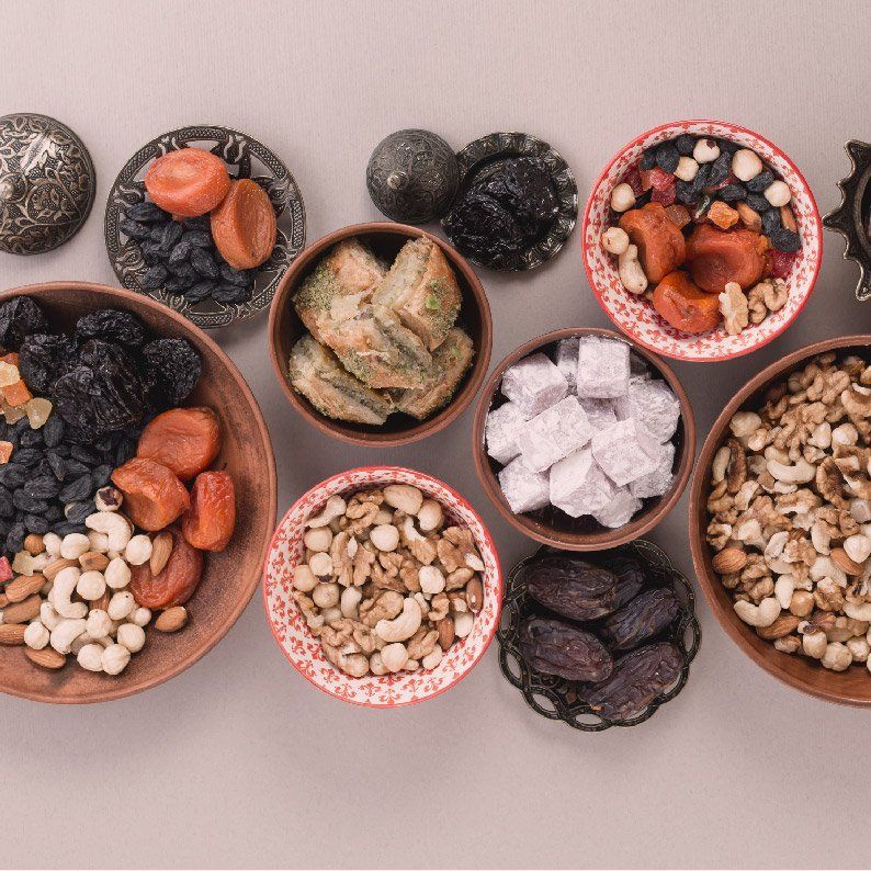 Variedad de frutos secos, semillas, frutas deshidratadas y cubos de coco en pequeños cuencos sobre una superficie beige.
