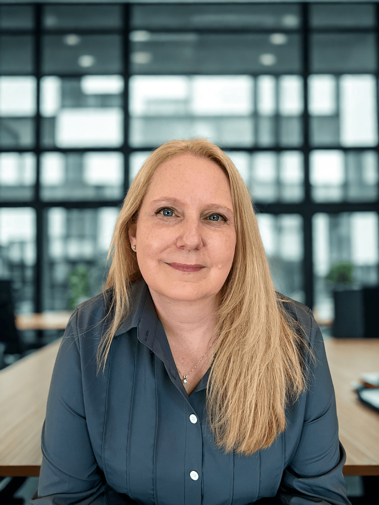 Woman with long blonde hair, smiling in a modern office, wearing a dark blue shirt.