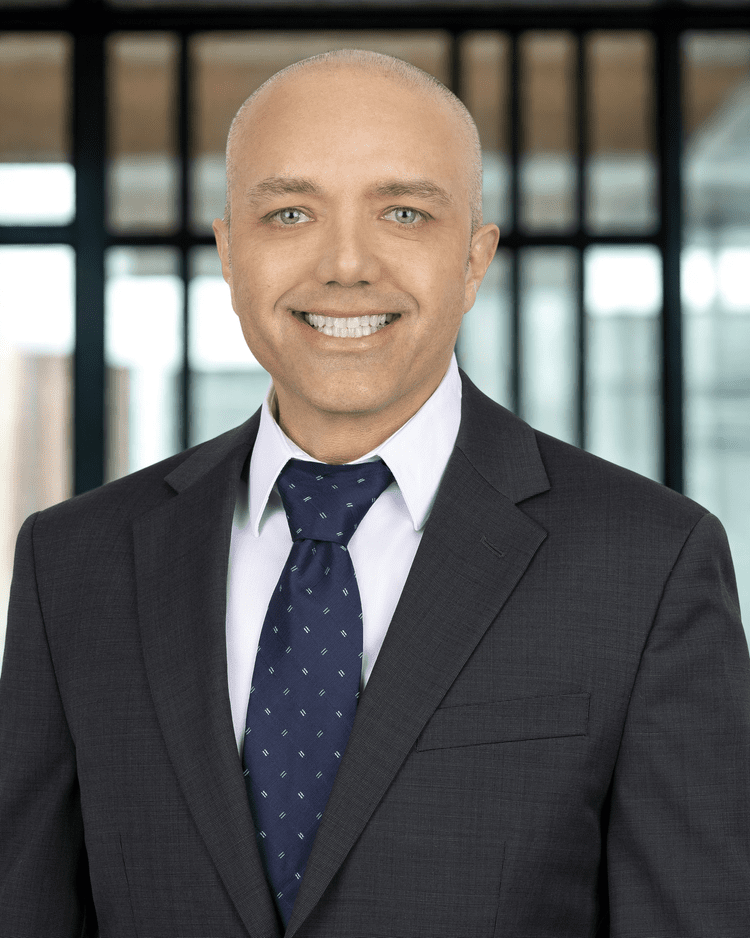 Man in a suit, smiling, in front of a window. He has short hair and a blue patterned tie.