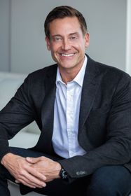 Man in blazer and open-collared shirt smiles while seated against a white background.