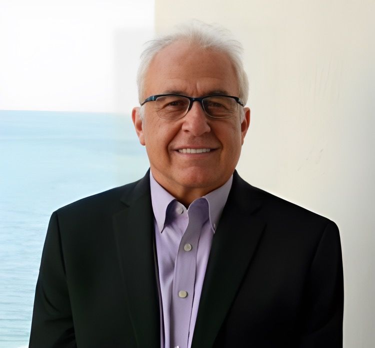 Man in suit and tie smiling. Background: blurred office setting.