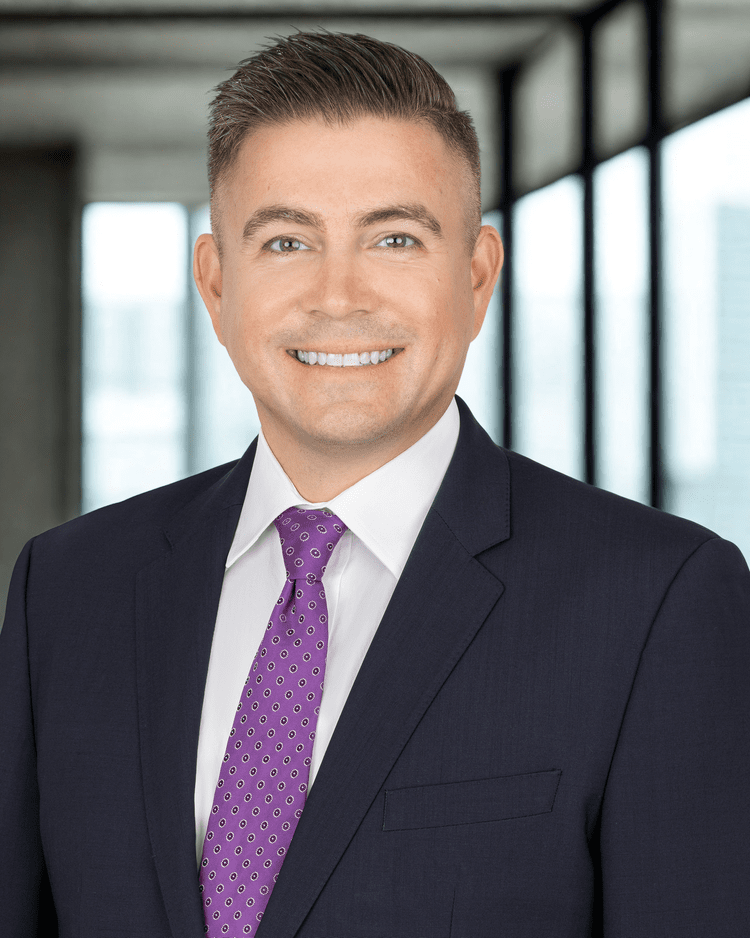 Man in a dark suit with purple tie smiles, posing in a modern office setting.