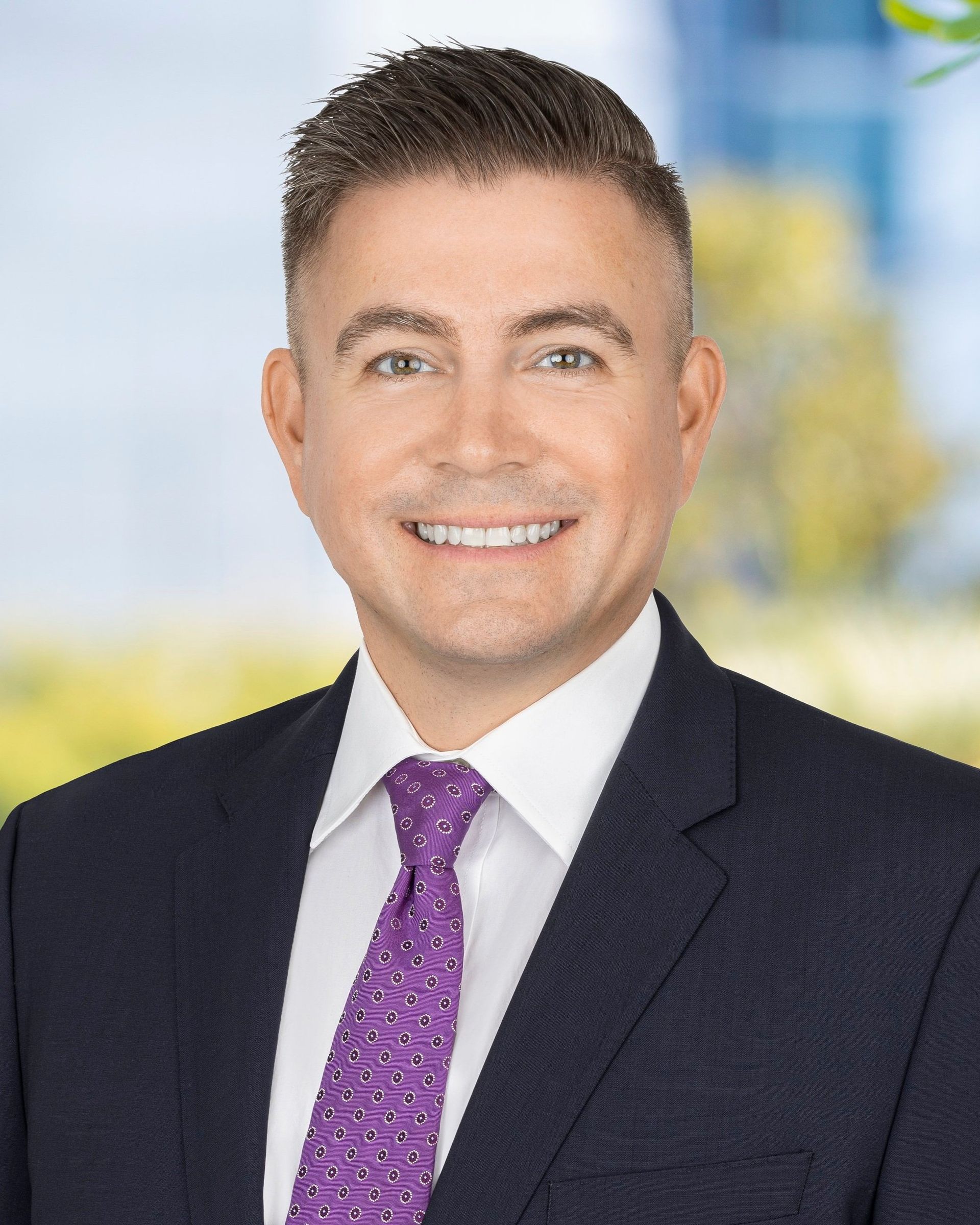 Man in suit and tie smiling. Background: blurred office setting.