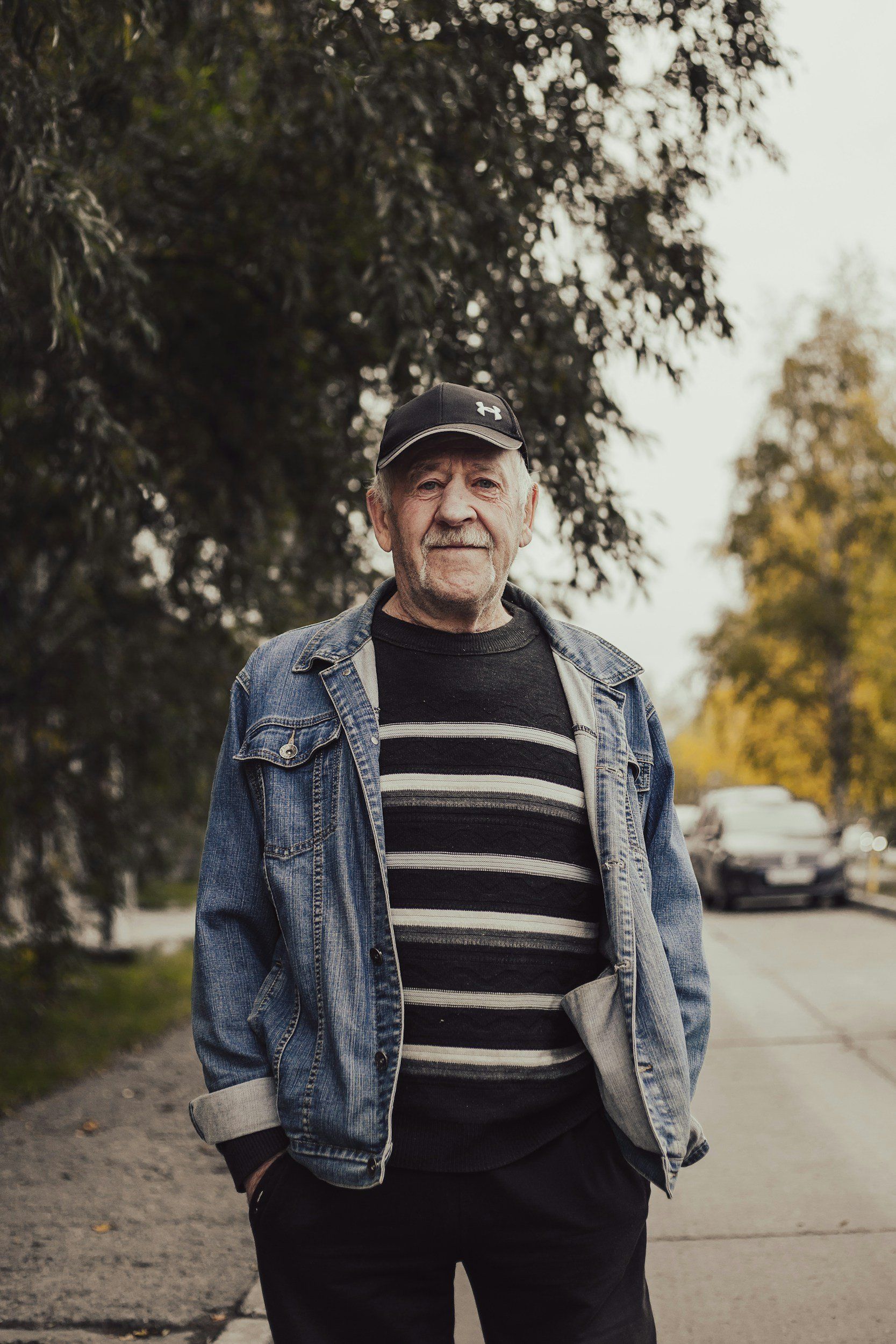 Man in denim jacket and striped sweater smiles on a path, trees and cars in the background.