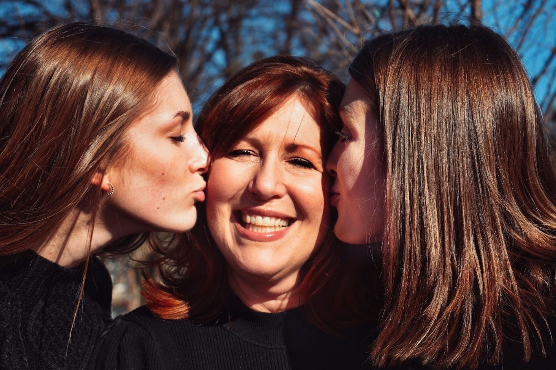 Woman smiles as two people kiss her cheeks outside.