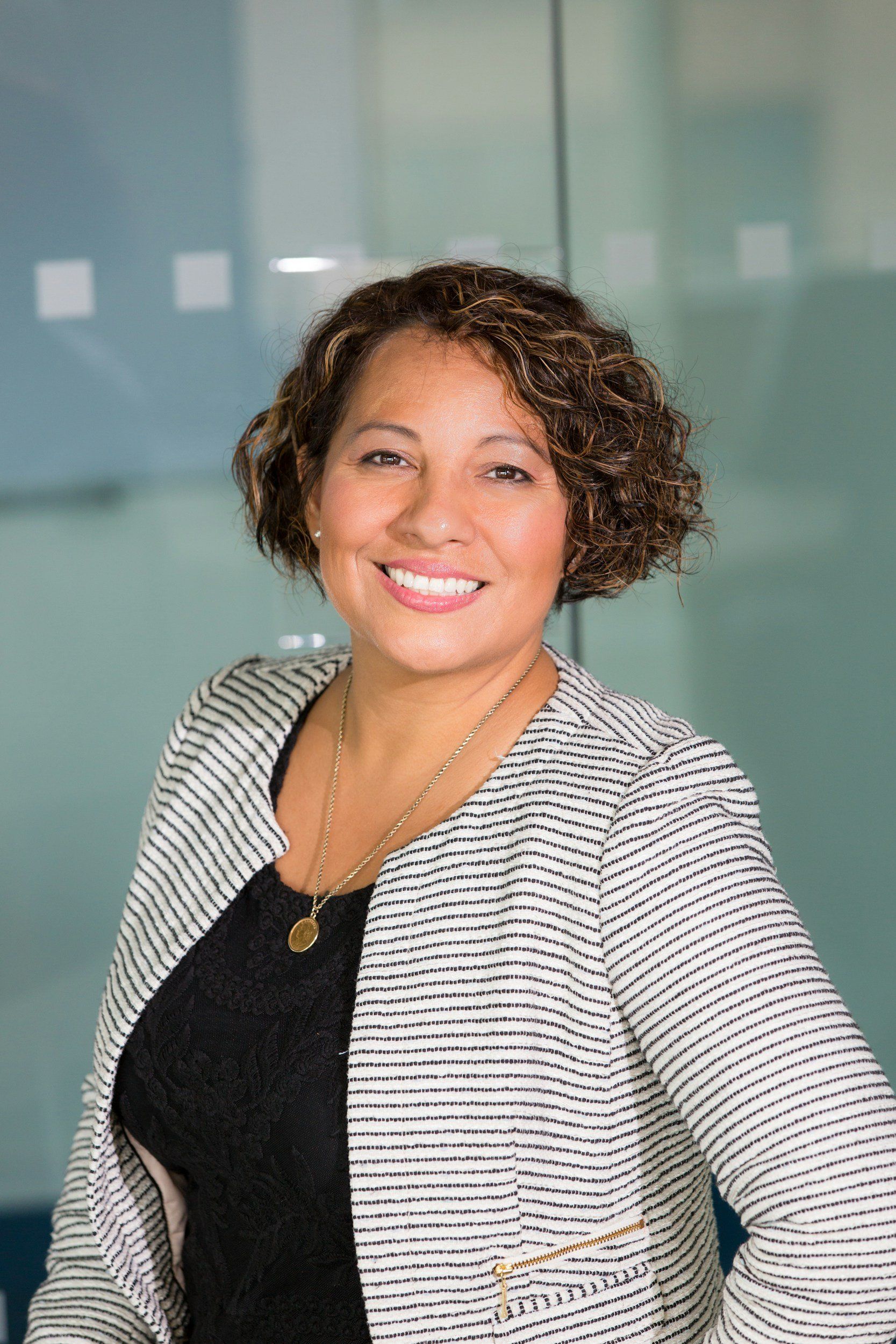 Woman with curly brown hair, smiling, wearing a black top and striped blazer, in front of a glass wall.
