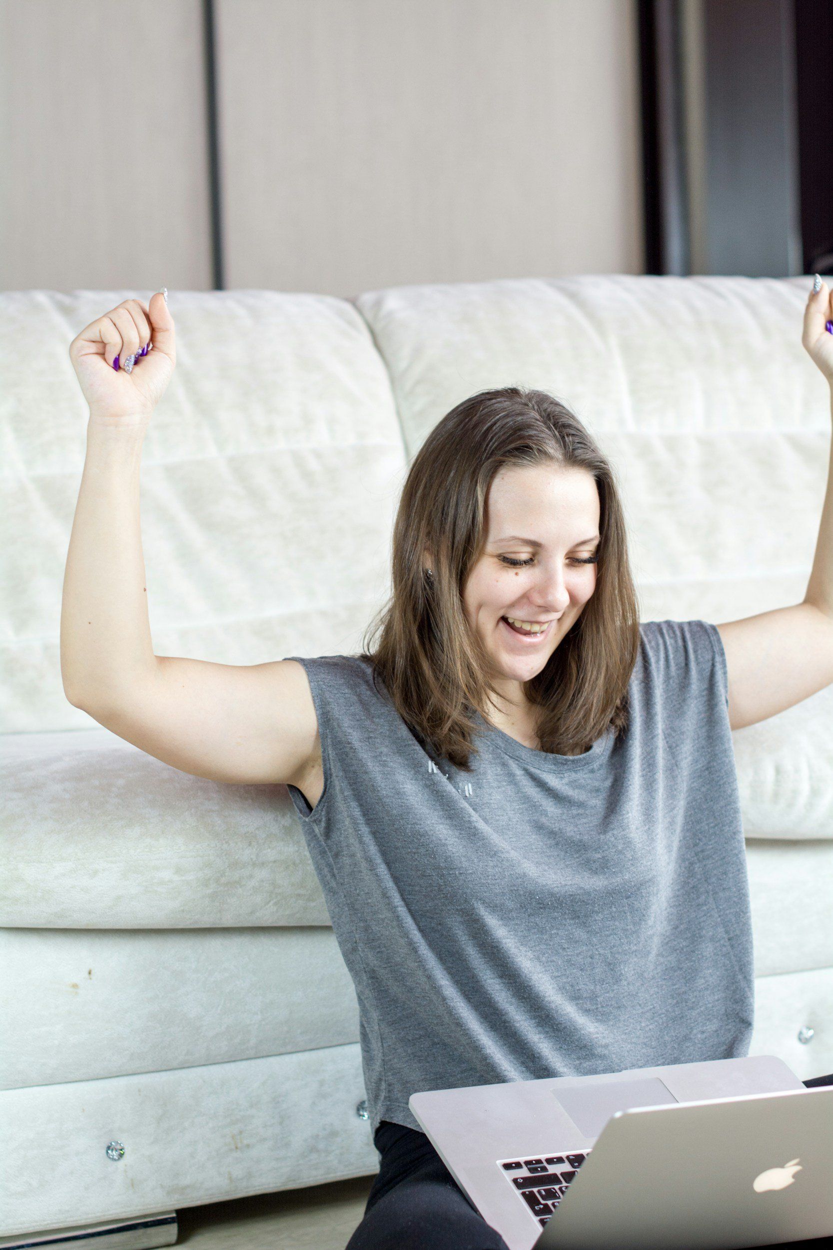 Woman seated on floor with arms raised, smiling at laptop in front of a white couch.