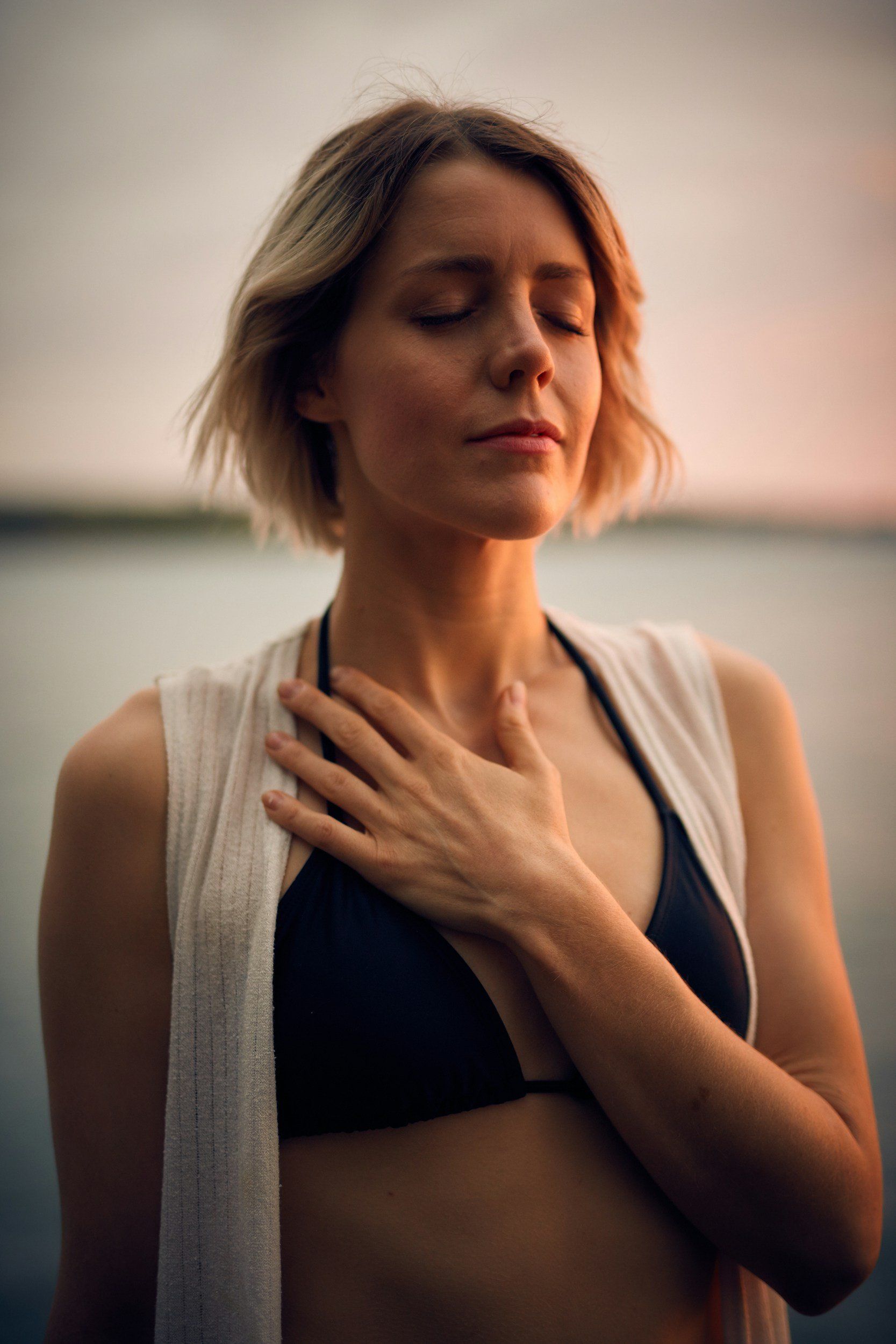 Woman with eyes closed, hand on chest, wearing a black top and white vest, outdoors near water.