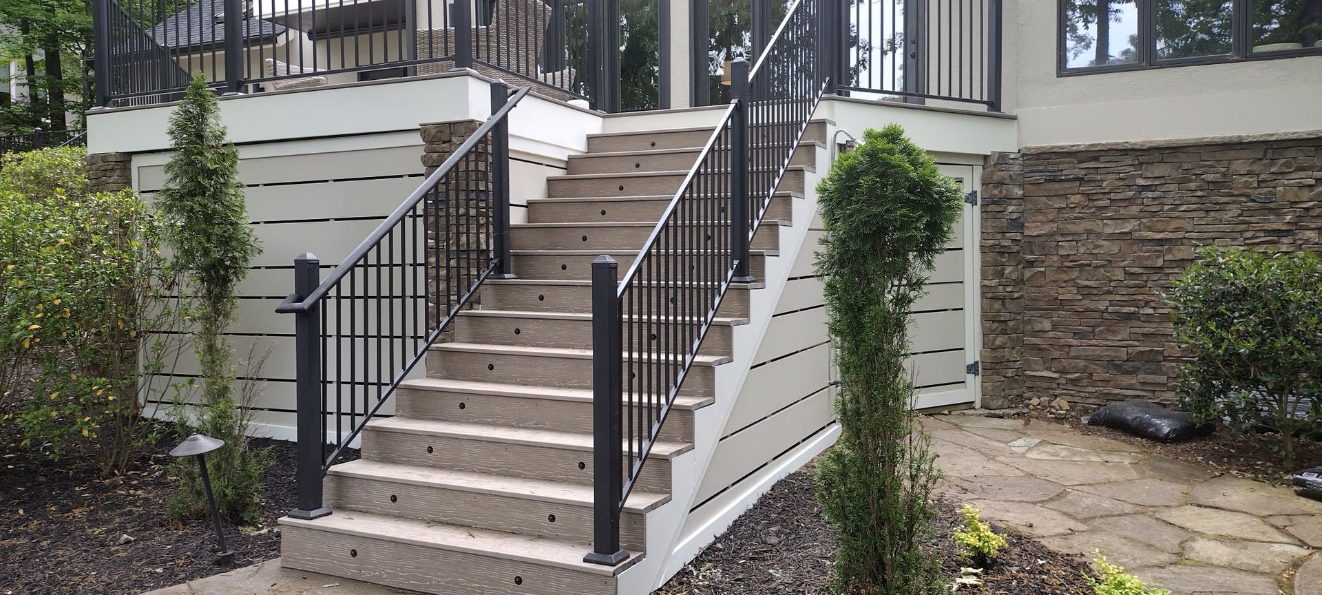 Exterior stairs with black railing leading to a raised deck. Light-colored siding and stone wall.