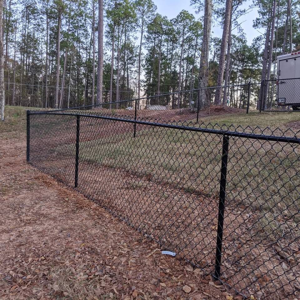 A chain link fence surrounds a dirt field with trees in the background.