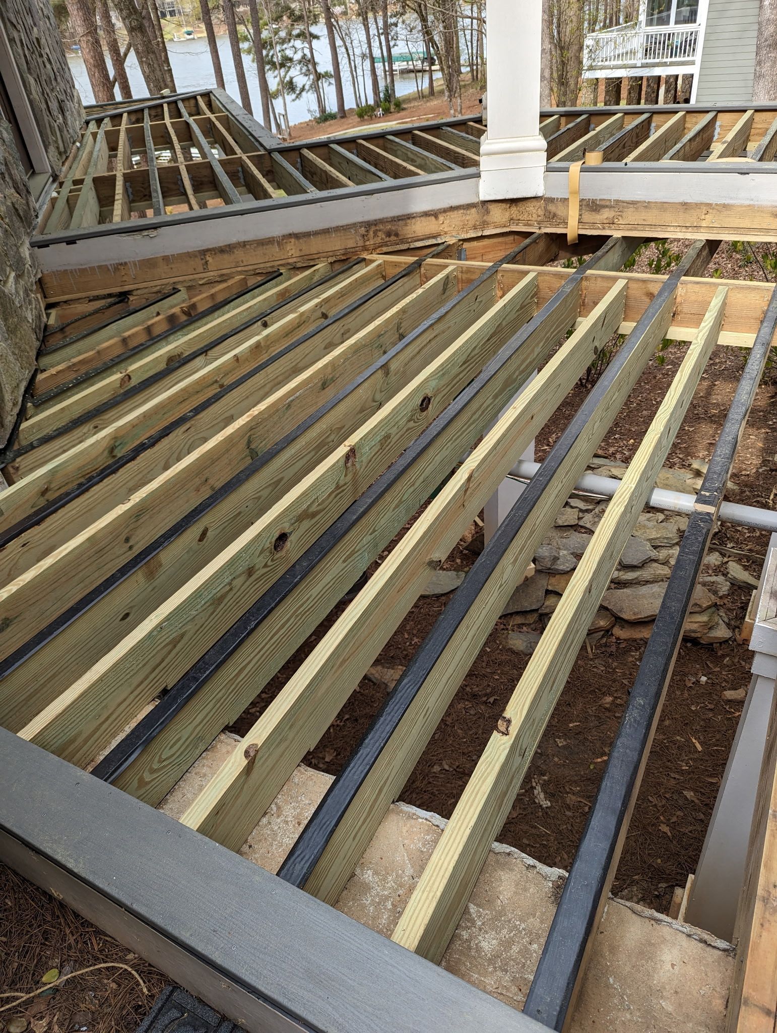 Deck under construction: wood frame with dark strips between boards, partial view of lake.
