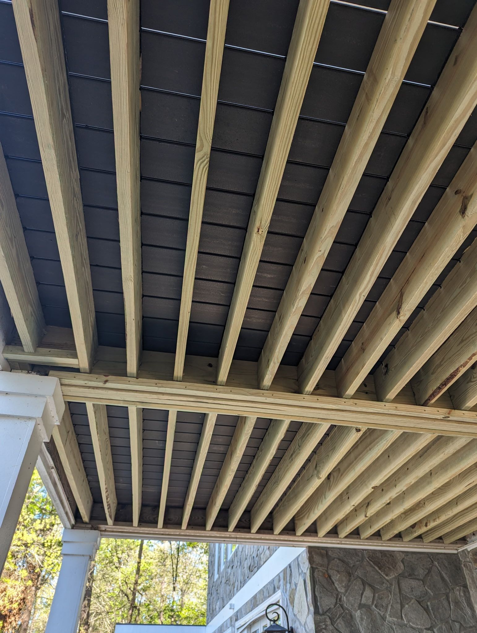 Underside view of a wooden deck. The deck has horizontal beams and a black underlayment.