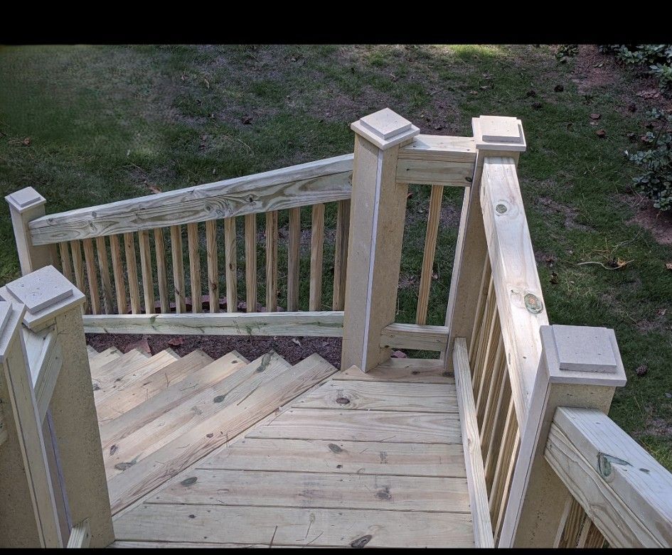 Wooden deck with stairs, railing, and square posts. The deck has a natural wood tone, and it's surrounded by green grass.