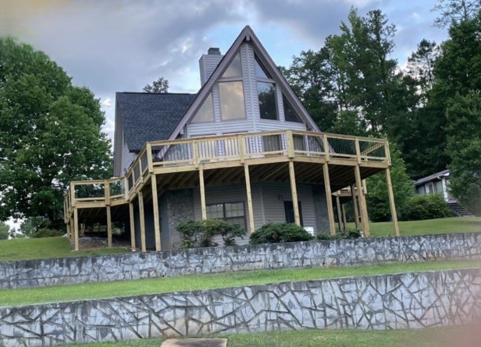 A-frame house with a wraparound wooden deck, set on a grassy hill with a stone retaining wall.