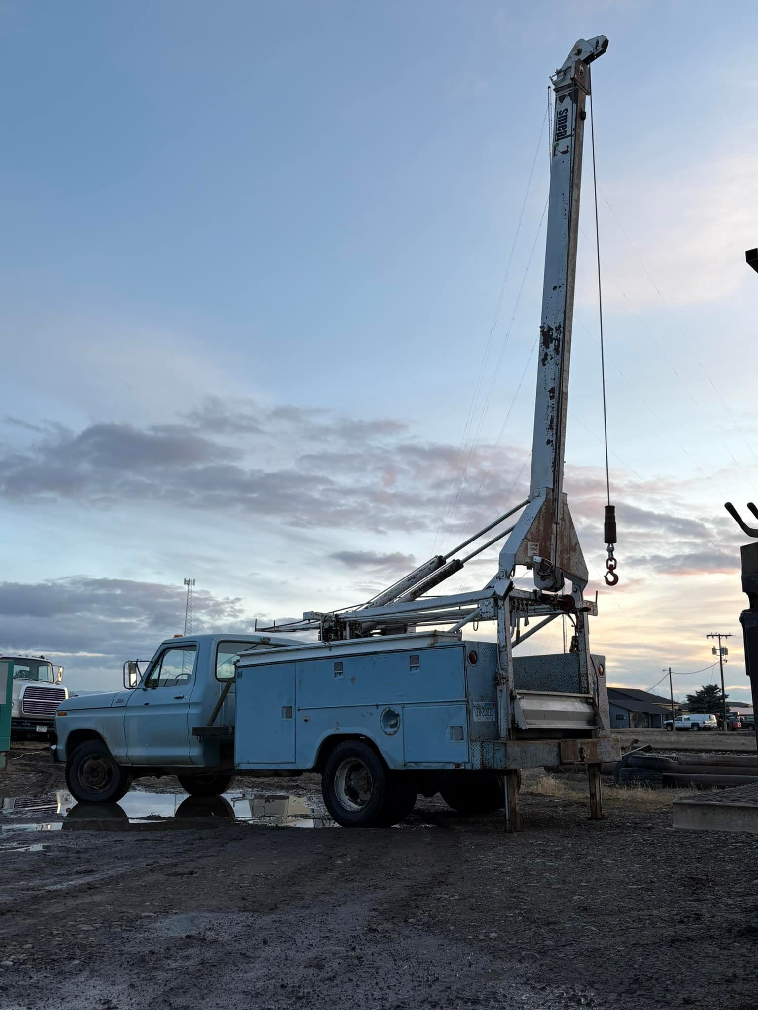 Light blue well drilling truck with tall drilling equipment, parked outside, against a cloudy sky.