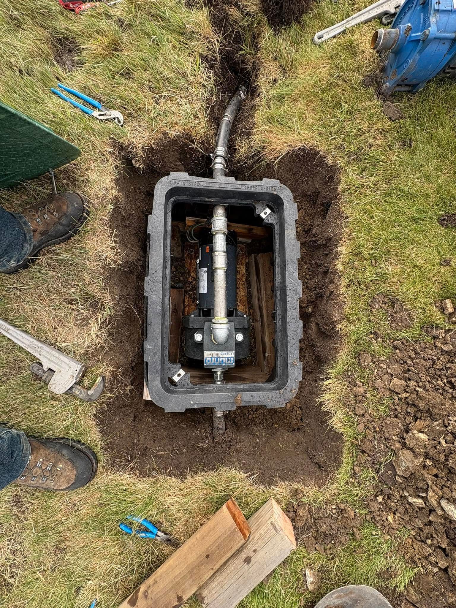Open irrigation valve box in a grassy yard, with tools and a partial view of boots.