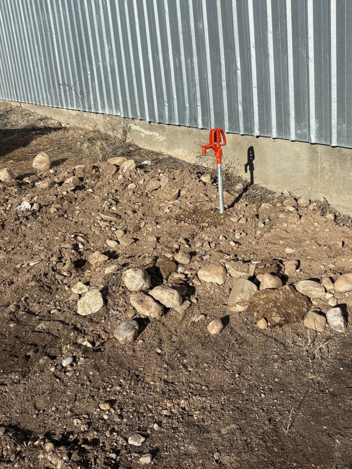 Orange water pump next to a corrugated metal wall, surrounded by dirt and rocks.