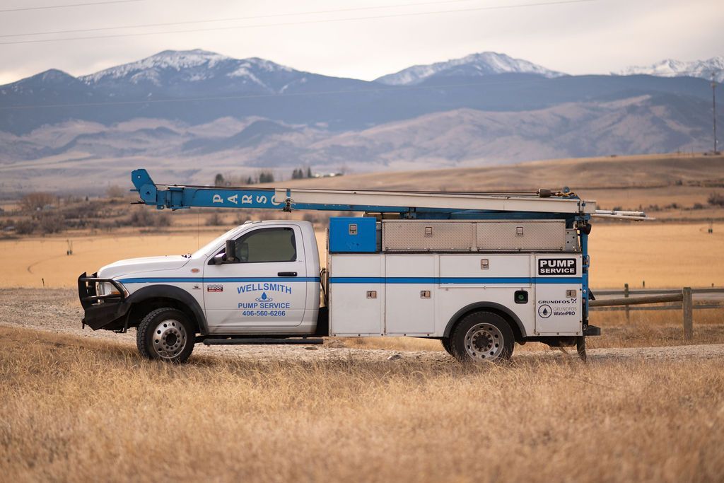 A white utility truck with a blue boom lift parked in a dry, grassy field with snow-capped mountains in the background.