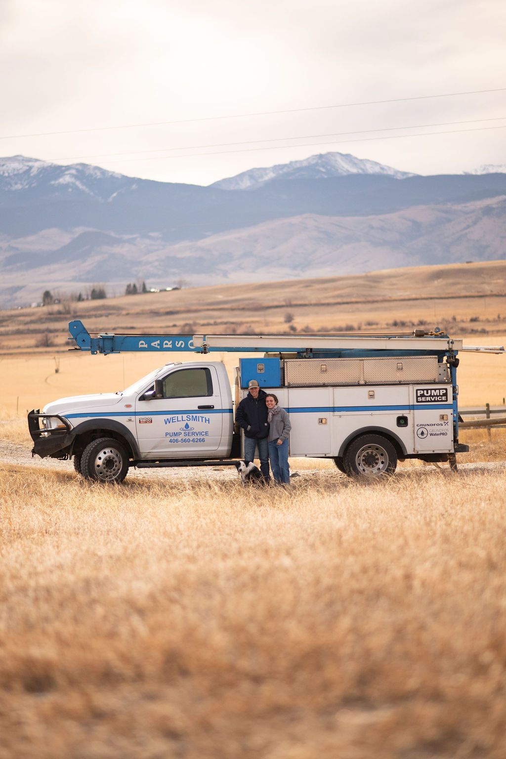 Old blue truck with drilling rig in a forest, person near white pipes.