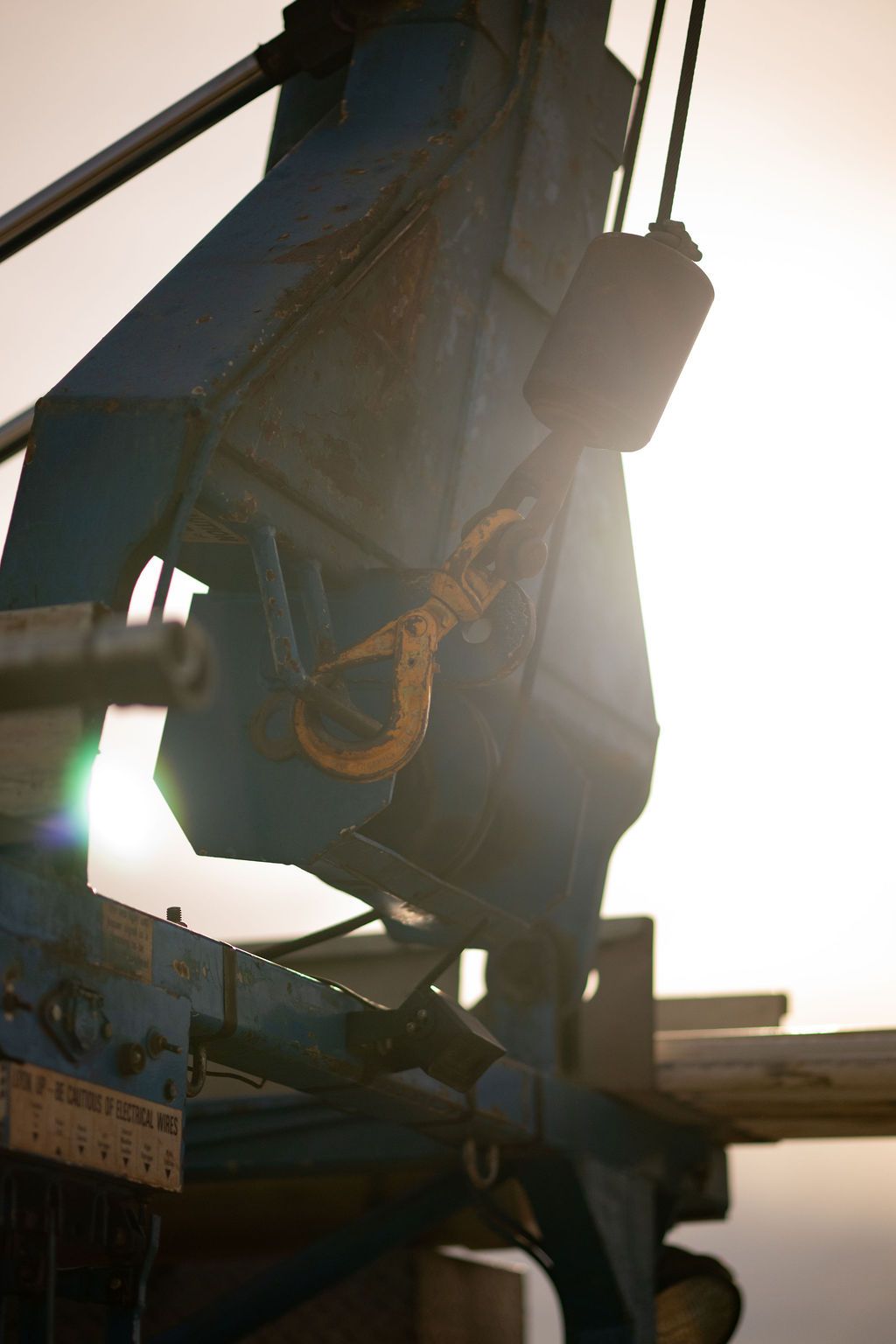 A close-up of a blue industrial crane hook and pulley system silhouetted against a bright, hazy sky.