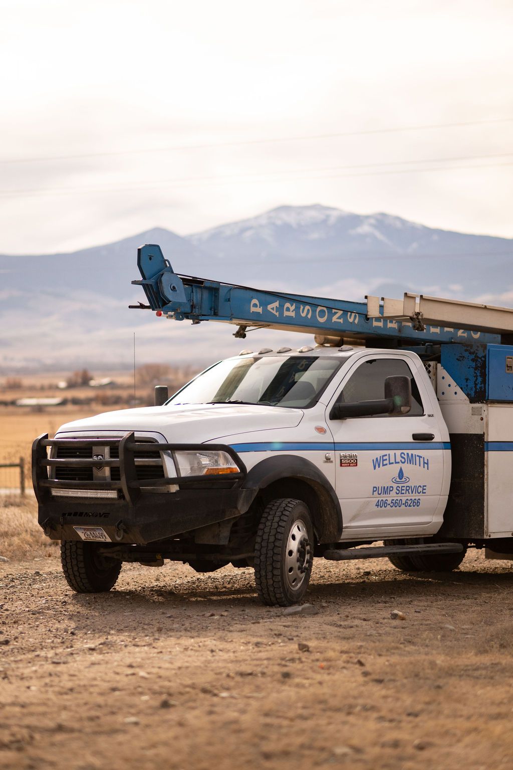 A white utility truck with a blue crane boom parked on a dirt lot with a snow-capped mountain in the background.
