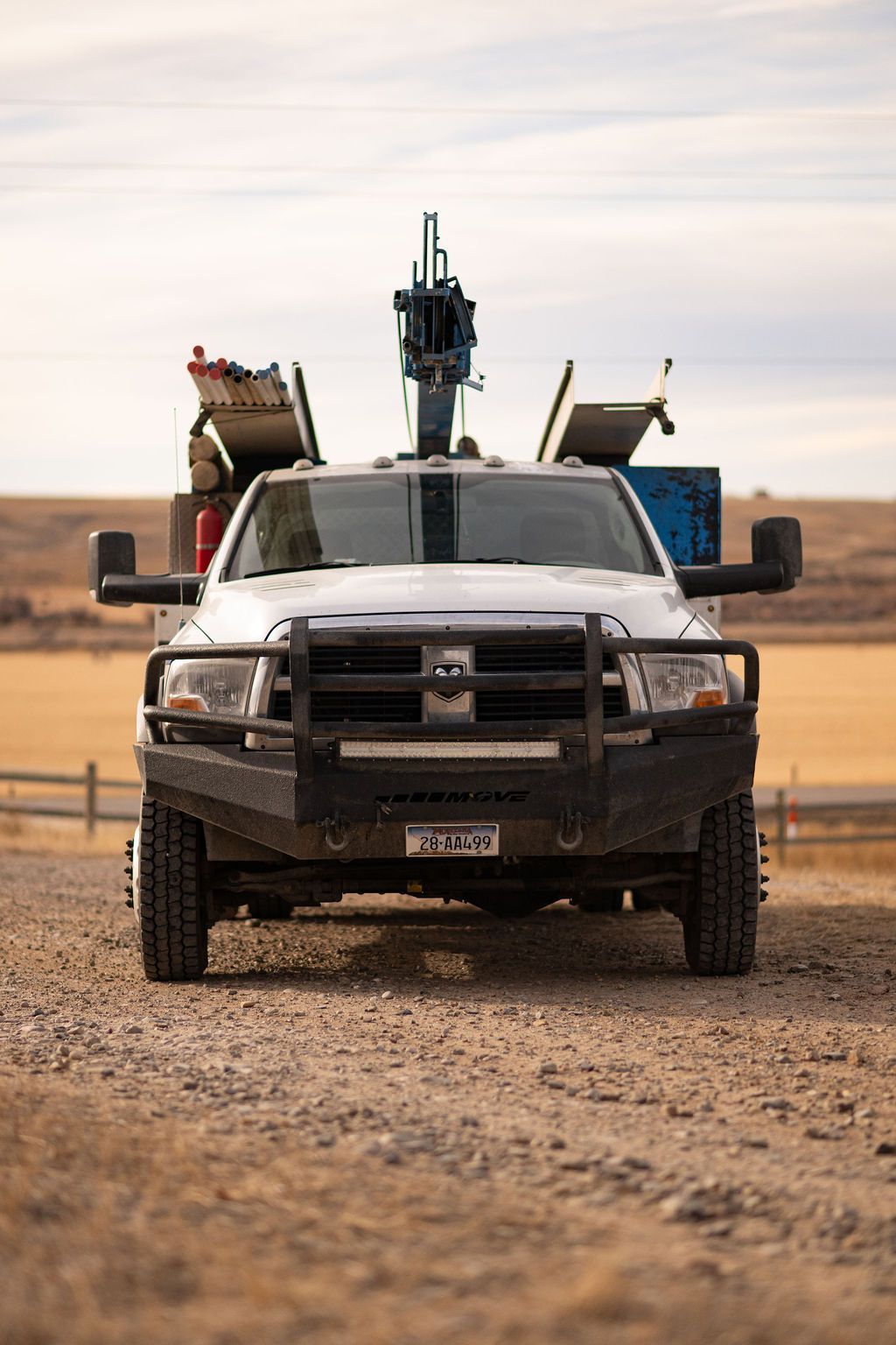 Front view of a white utility truck with a crane attachment, parked on a gravel road in a rural, open field setting.