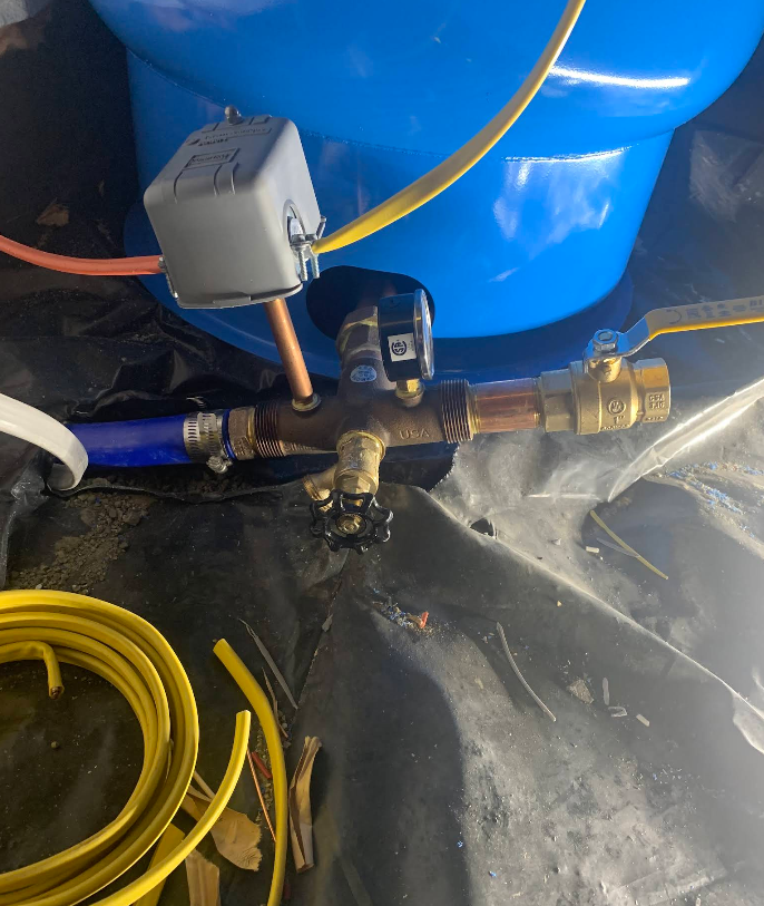 Well pump components, including pressure switch, pipes, and valves, against a blue tank and black tarp.
