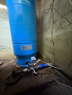 Blue water pressure tank with attached pipes and gauge in a concrete room.