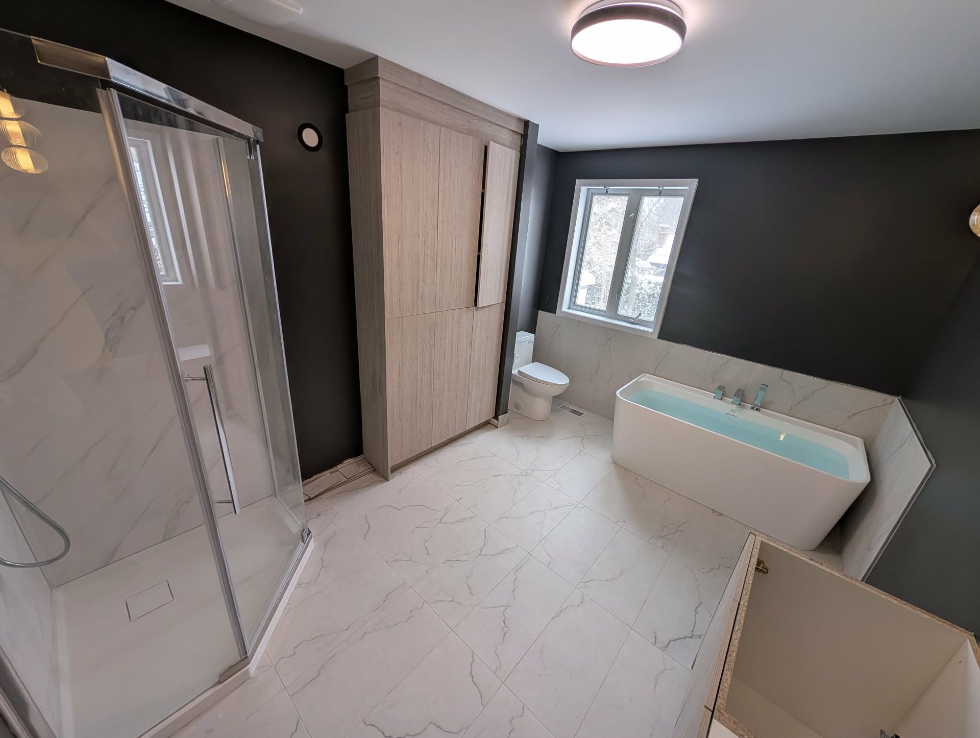 Salle de bains moderne avec douche, baignoire, toilettes et grand placard ; murs gris foncé, robinetterie blanche, placard en bois clair.