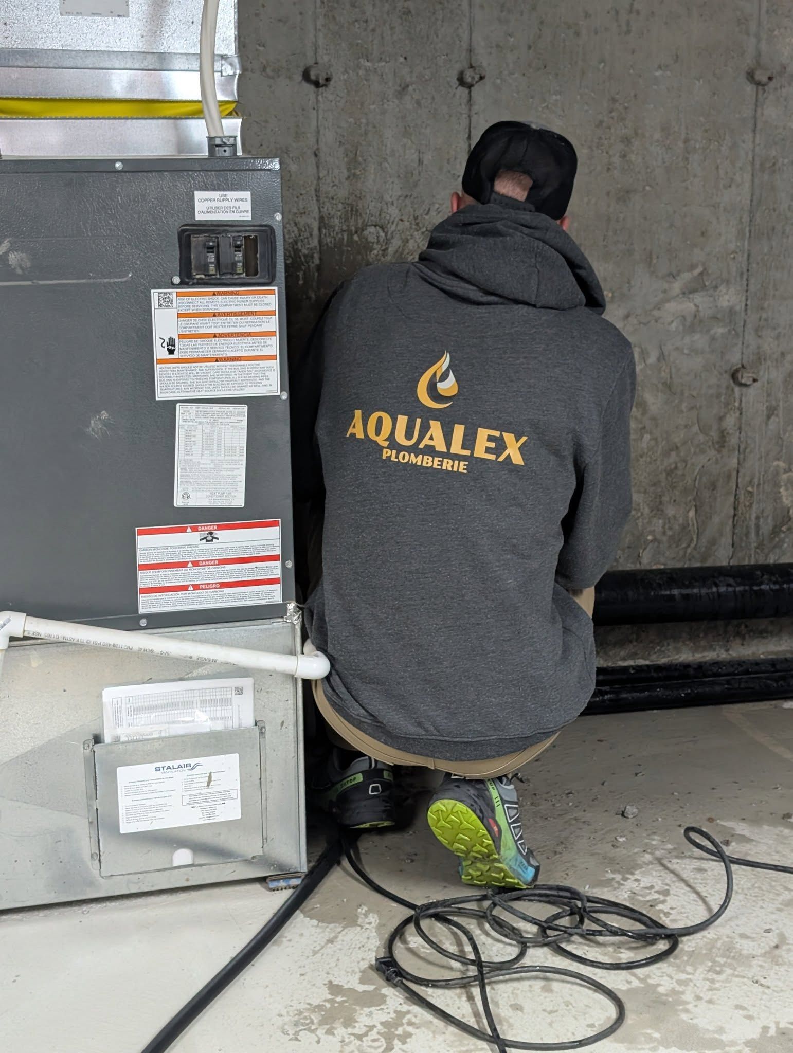 Une personne travaille sur un système de climatisation dans un sous-sol. Elle porte un sweat-shirt gris et est accroupie près de l'appareil. Un câble noir est posé au sol.